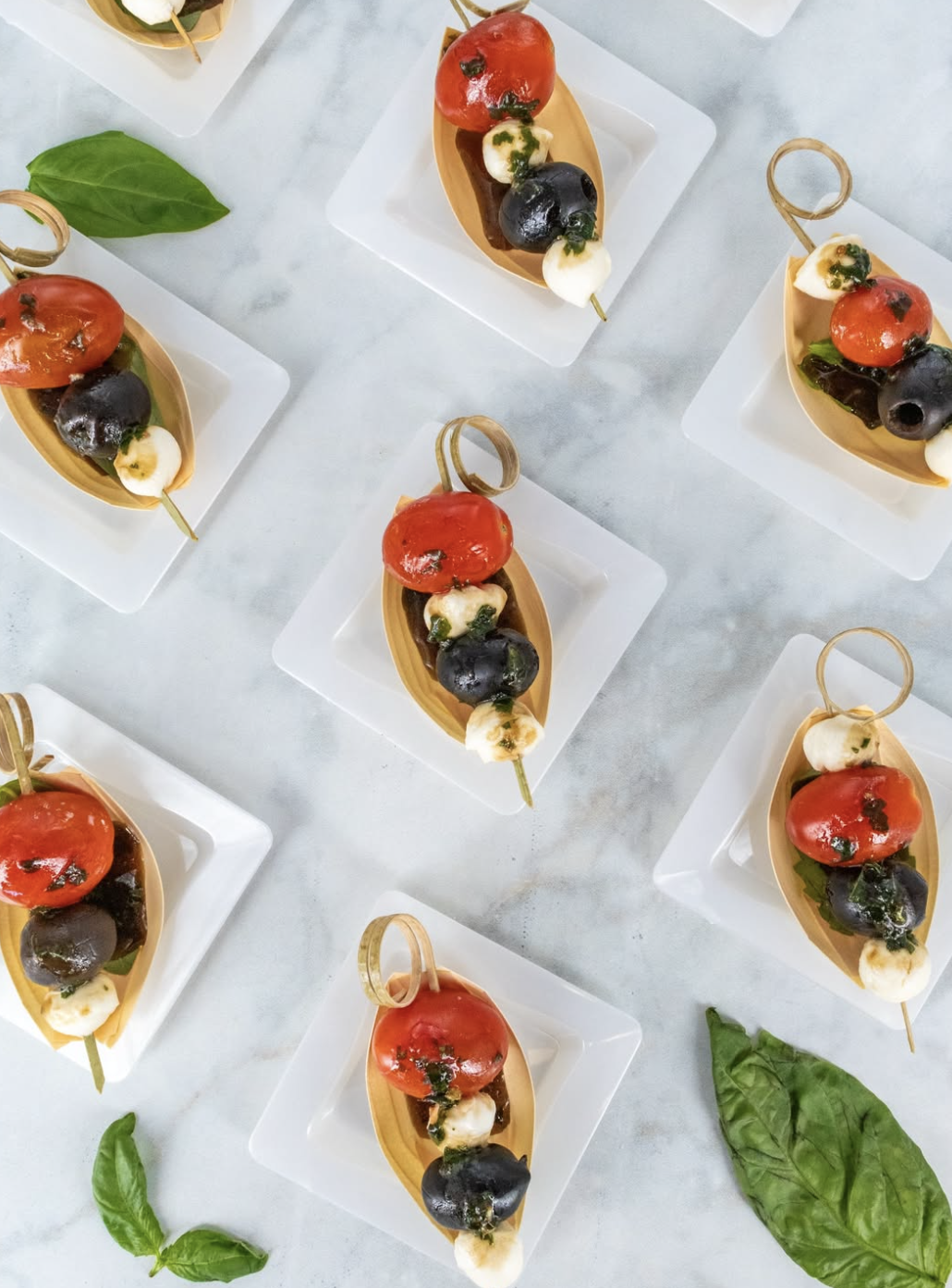 Multiple servings of Caprese salad skewers with cherry tomatoes, mozzarella balls, basil leaves, and balsamic glaze on small plates with green basil leaves scattered around.