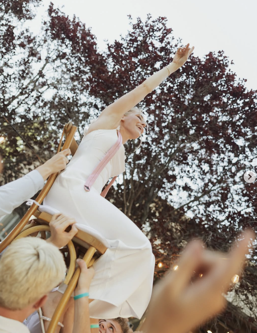 A woman with blonde hair, wearing a white dress and a pink ribbon, is sitting on a wooden chair being carried on people's shoulders. She is smiling and reaching upward with her right arm, in an outdoor setting with trees in the background.