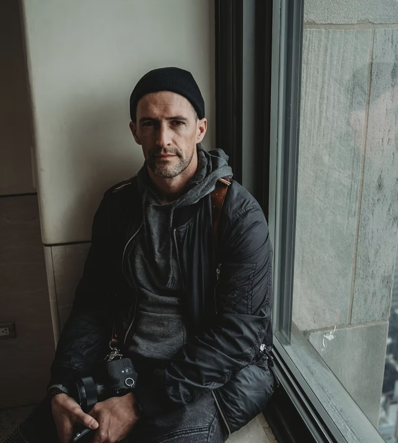 A man sitting by a window, holding a camera, wearing a black beanie, a black jacket, and a gray hoodie inside.