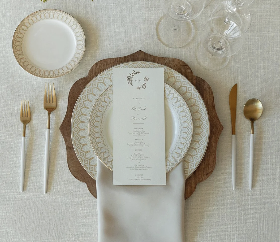 Elegant wedding table setting with gold and white dinnerware, gold cutlery, a white napkin, empty wine glasses, and a wedding menu placed on a wooden charger