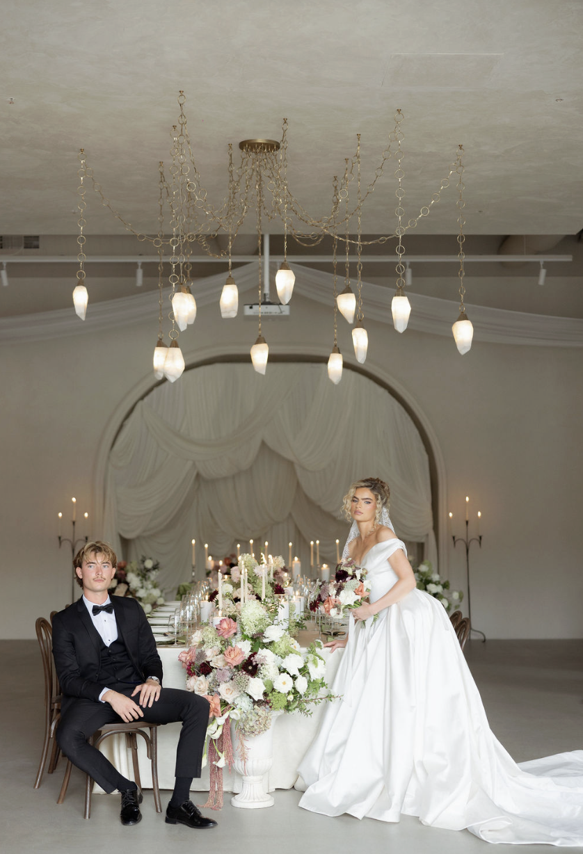 A bride in a white wedding dress holding a bouquet and a groom in a black tuxedo seated in a beautifully decorated wedding reception hall with floral arrangements, candles, and elegant drapery.