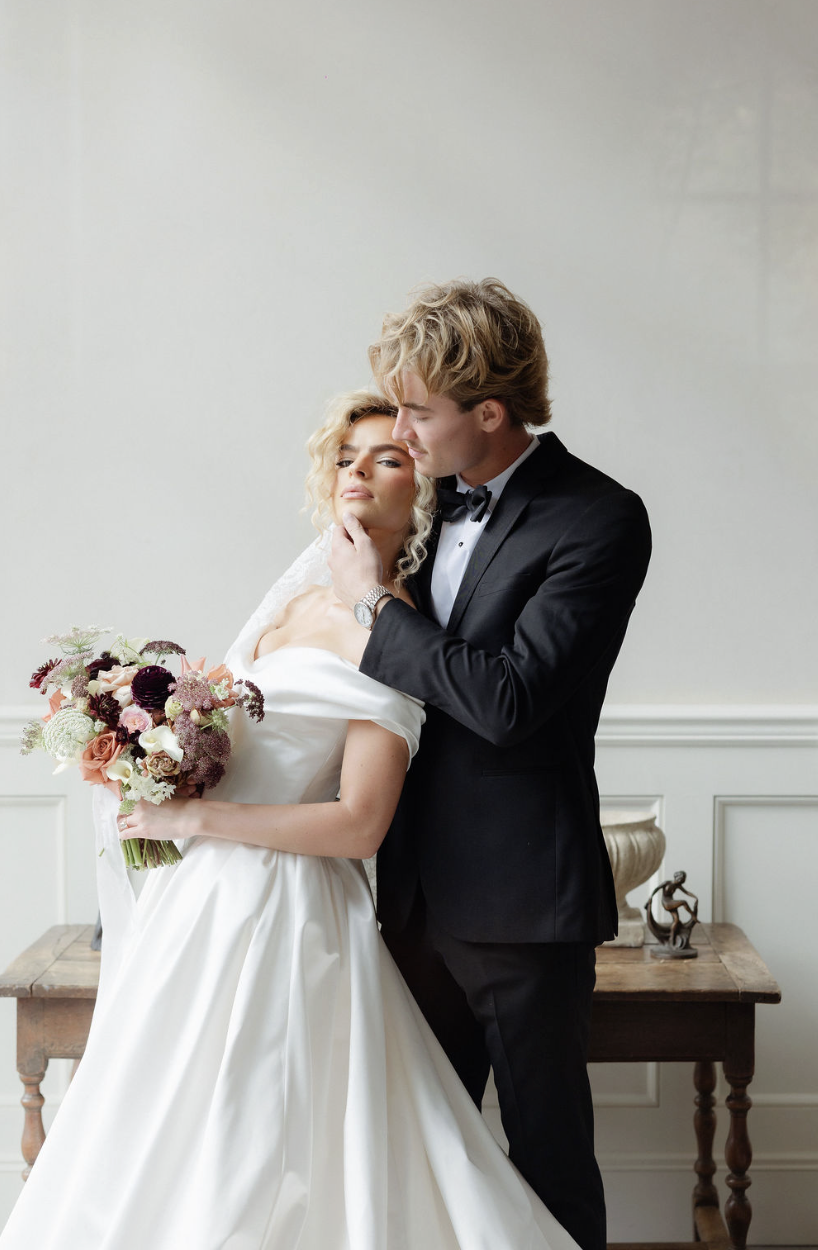A bride and groom in wedding attire posing indoors. The bride is holding a bouquet of flowers and looking confidently at the camera, while the groom is gently touching her chin and looking at her affectionately.