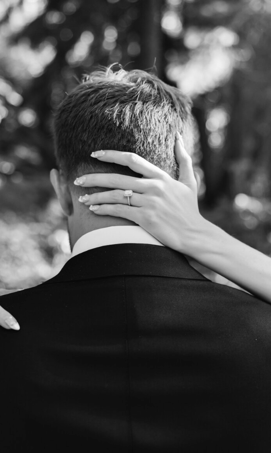 A woman with manicured nails and a ring on her finger hugs a man in a suit from behind, holding his head with one hand. The photo is in black and white, with trees blurry in the background.
