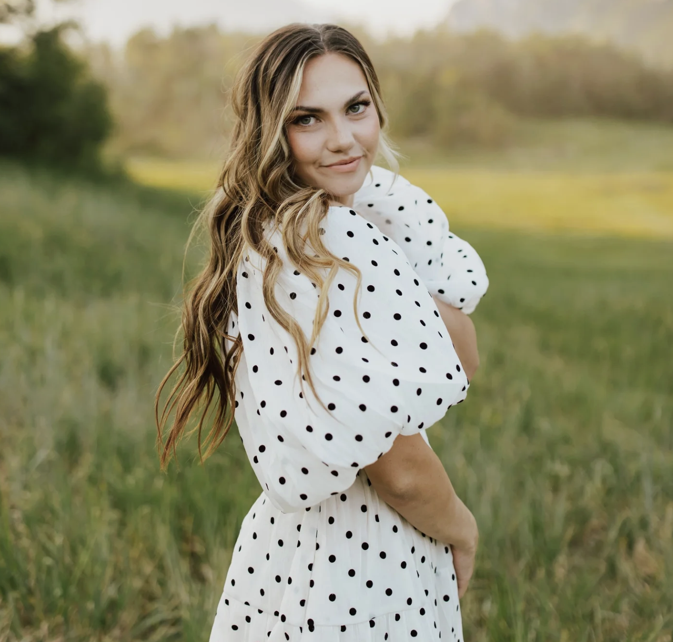 A young woman with long wavy blonde hair, wearing a white dress with black polka dots, standing outdoors in a grassy field during golden hour.