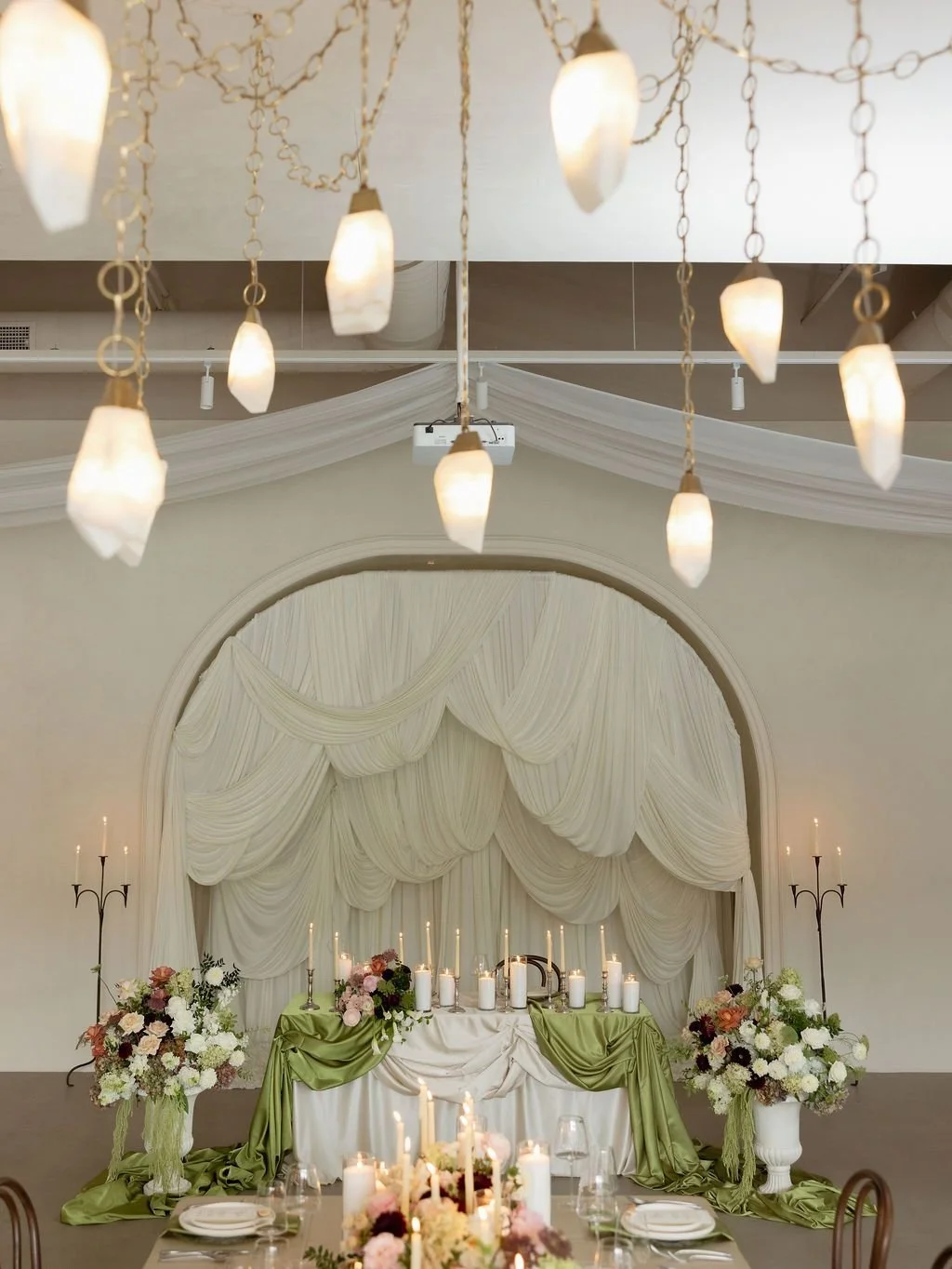 Elegant event space decorated with draped white fabric, floral arrangements, and lit candles, featuring a high table with white and green fabric accents and two tall candle holders.