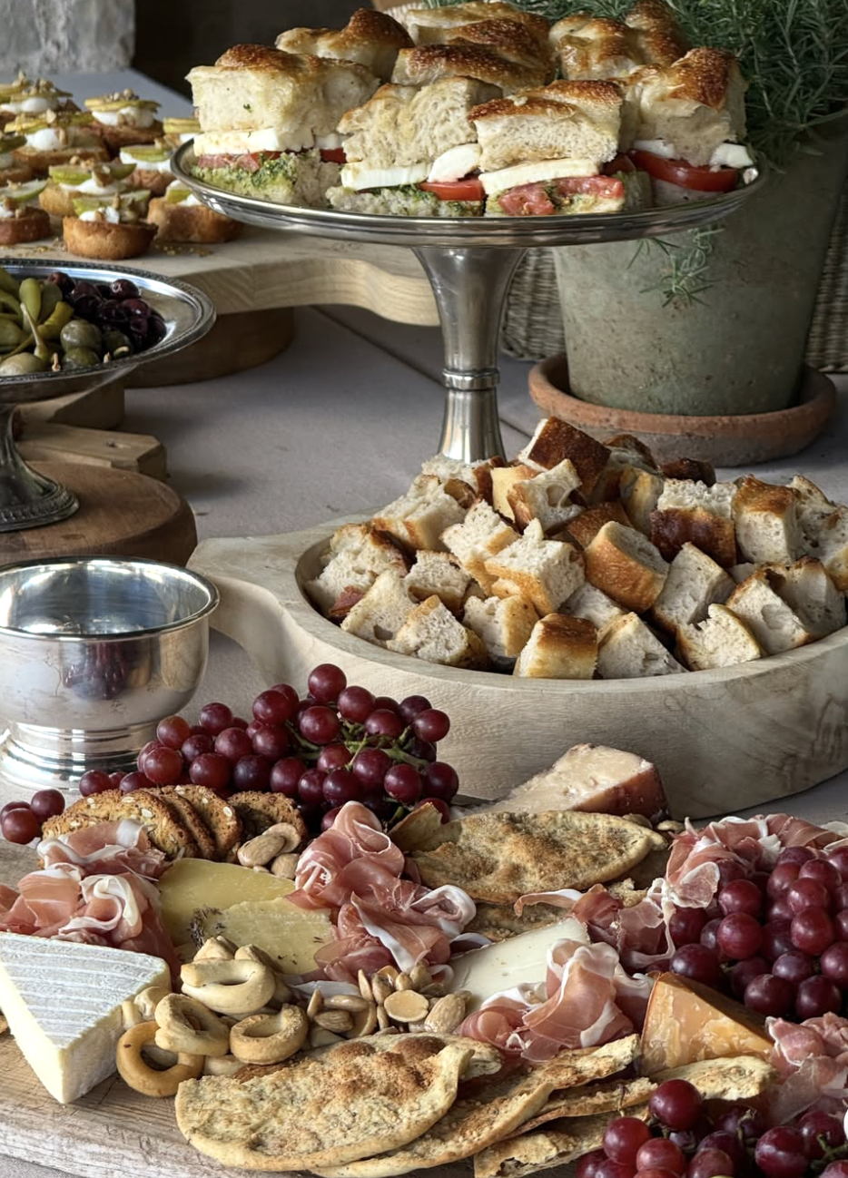 A spread of assorted appetizers including a cheese and fruit platter, bread cubes, pita chips, grapes, prosciutto, nuts, and various finger foods on serving platters and boards.
