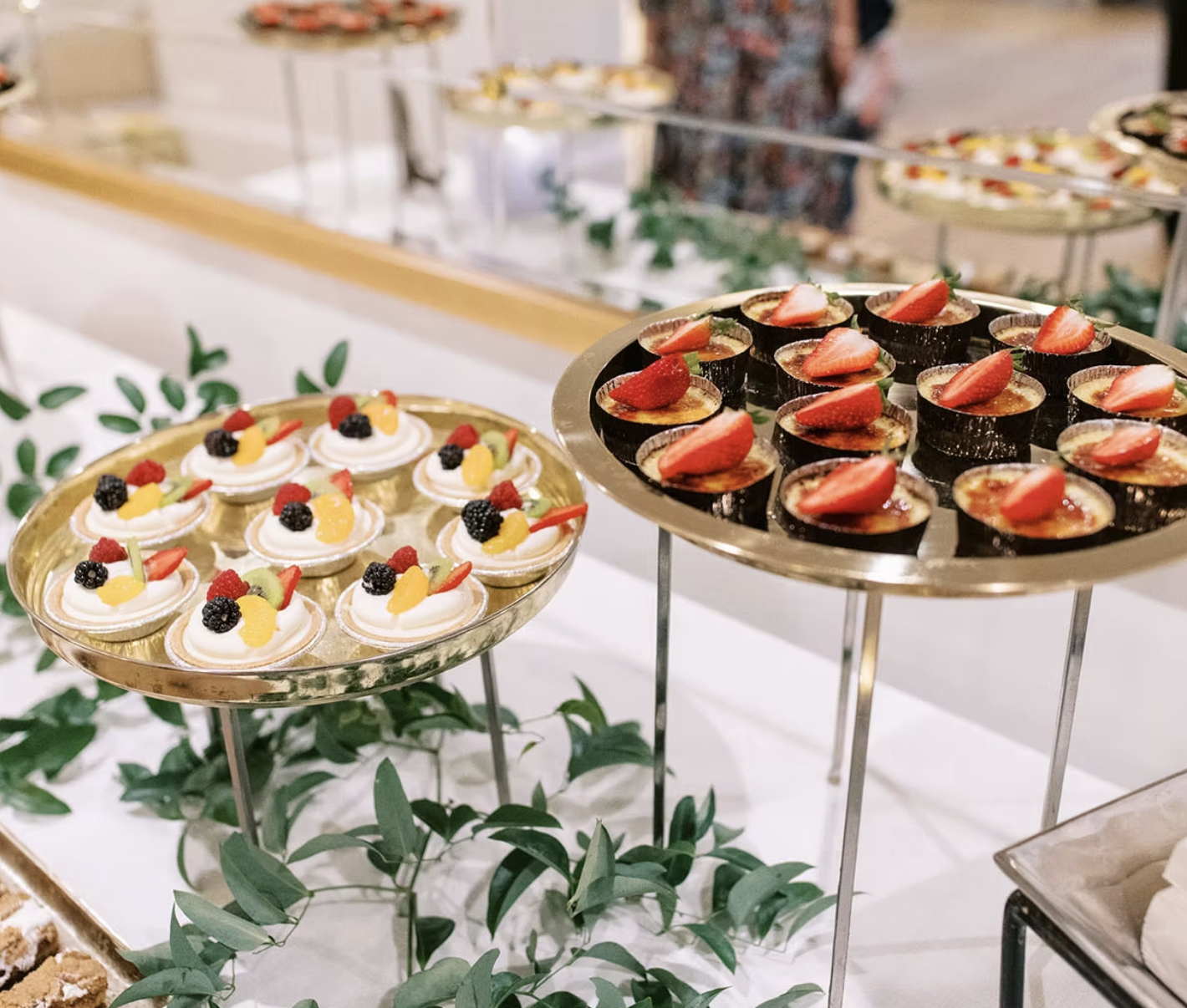 Assorted mini desserts on elegant serving trays, including fruit-topped tartlets and mini pavlovas with strawberries and blackberries, displayed on a white table with greenery.