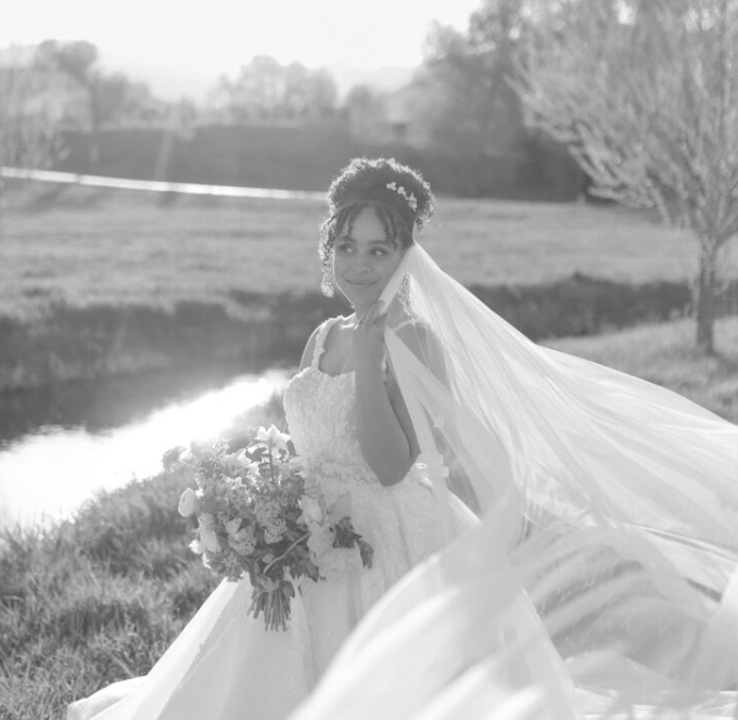 A bride standing outdoors near a river, holding a bouquet of flowers, wearing a wedding dress and veil, with trees and open field in the background during daylight.