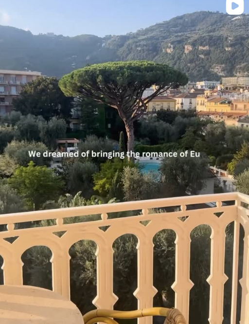 A scenic view from a balcony featuring a large, distinctive pine tree in the foreground, with a backdrop of residential buildings and green hills under a clear sky.