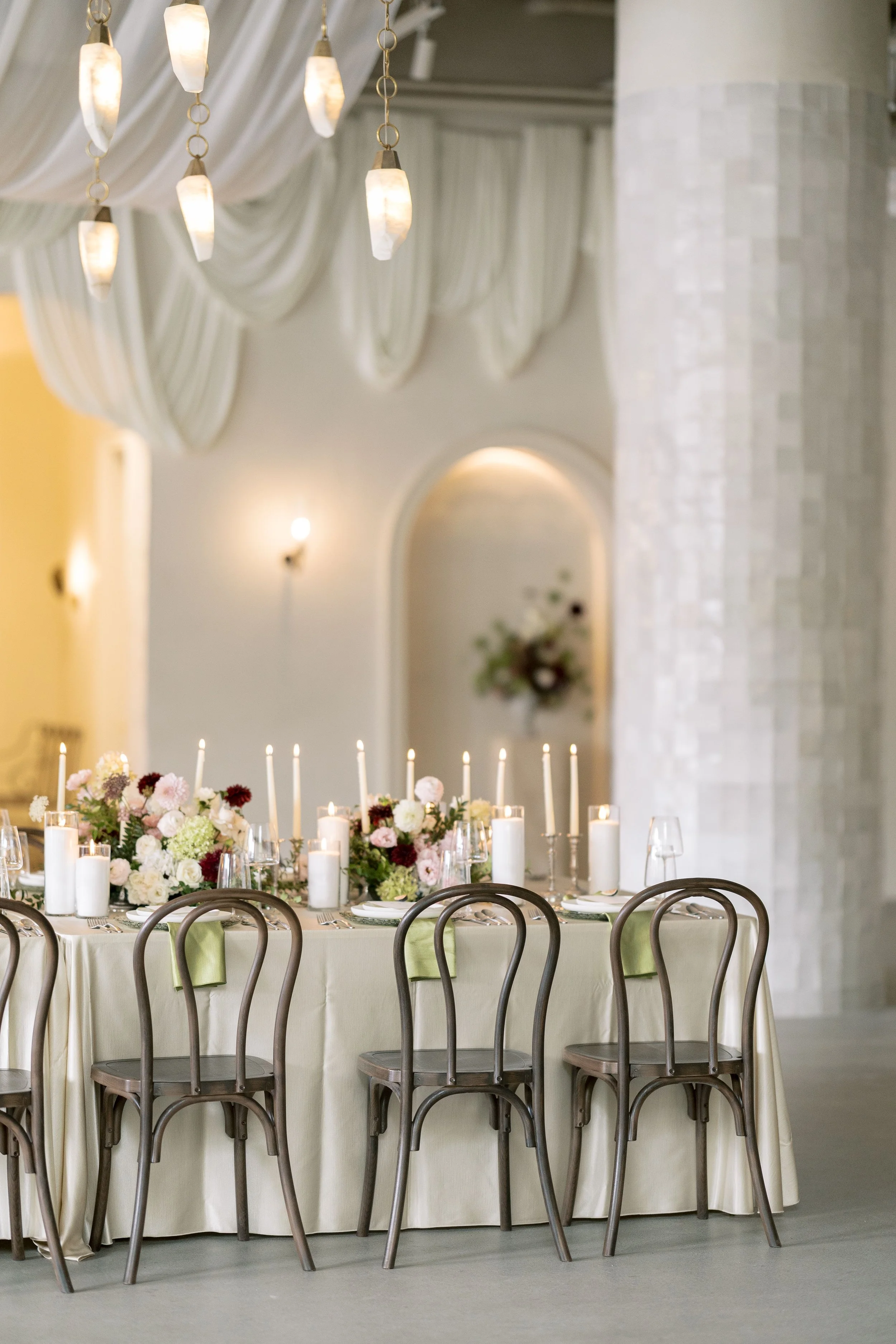 Round banquet table with a cream-colored tablecloth, decorated with a floral centerpiece and candles. Surrounding the table are wooden chairs with curved backs. The background features elegant, draped white fabric on the ceiling and soft lighting in an upscale event space.