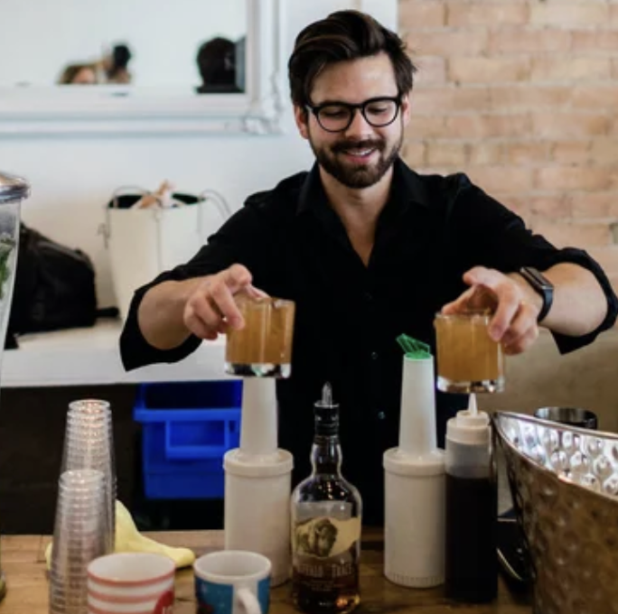 A man with glasses and a beard smiling while pouring two drinks into glasses at a bar or cafe.