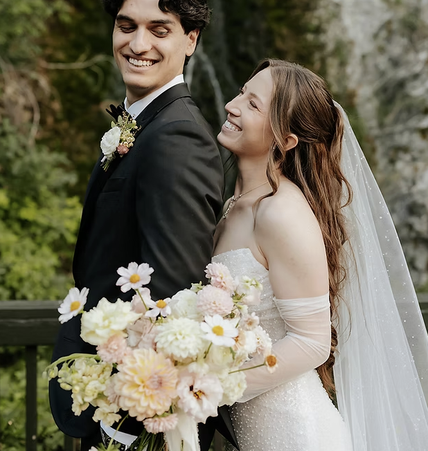 Bride and groom smiling, standing outdoors with trees, bride holding a large bouquet of flowers, wearing wedding dress and veil, groom in a suit with boutonniere.