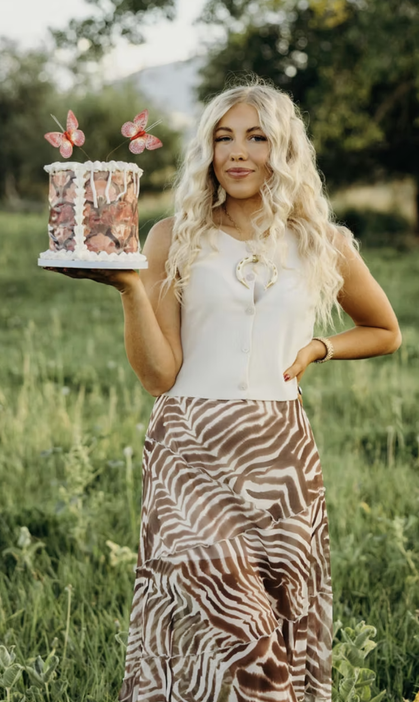 A woman with long, wavy blonde hair stands outdoors in a grassy area, holding a decorated cake with pink butterflies on top. She is wearing a white sleeveless blouse, a zebra-print skirt, and a bracelet, with a scenic background of trees and mountains.