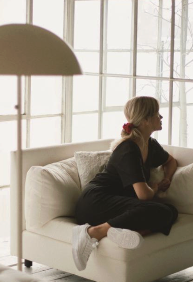 A woman with blonde hair tied with a red scrunchie, dressed in black, sits on a cream-colored sofa near a large window, looking outside.