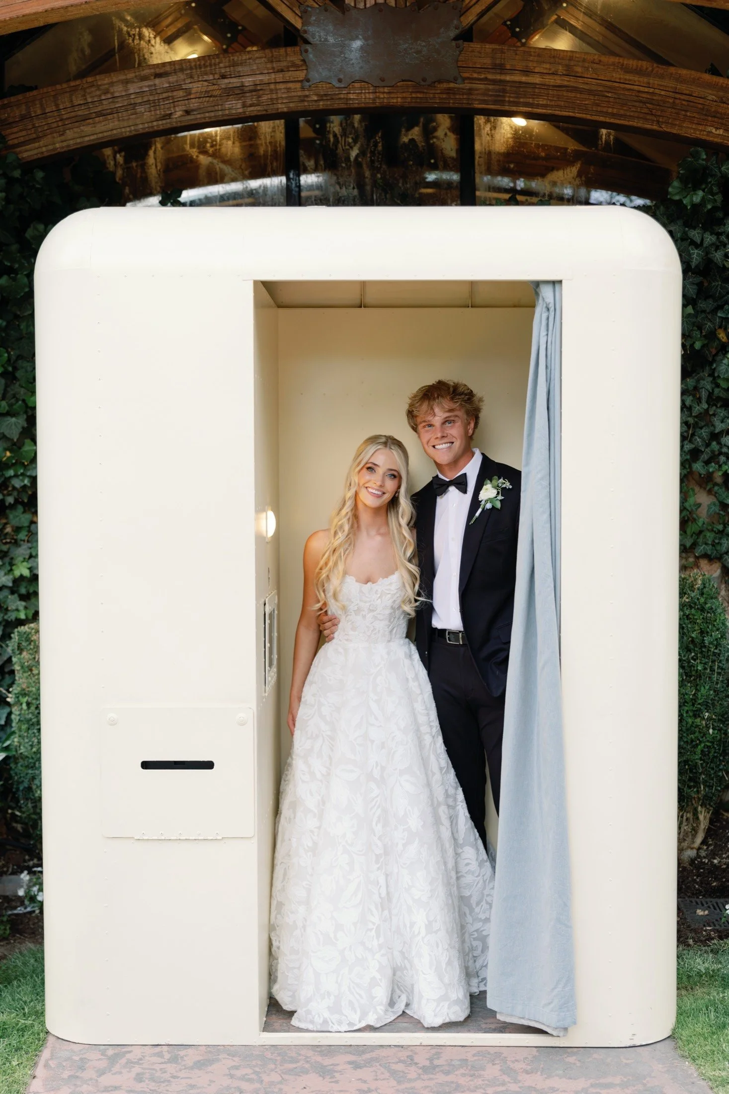 A bride and groom standing inside a small, cream-colored photo booth with gray curtains, outdoors on a lawn surrounded by green bushes