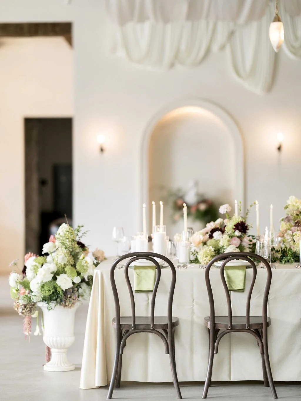 Elegant dining table setup with floral arrangements, candles, and glassware in a bright, sophisticated room with arched wall niche and wall sconces.