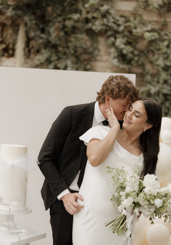 A couple sharing a kiss at their wedding, the bride holding a bouquet of white flowers, and the groom dressed in a black suit and white shirt, outdoors with greenery in the background.