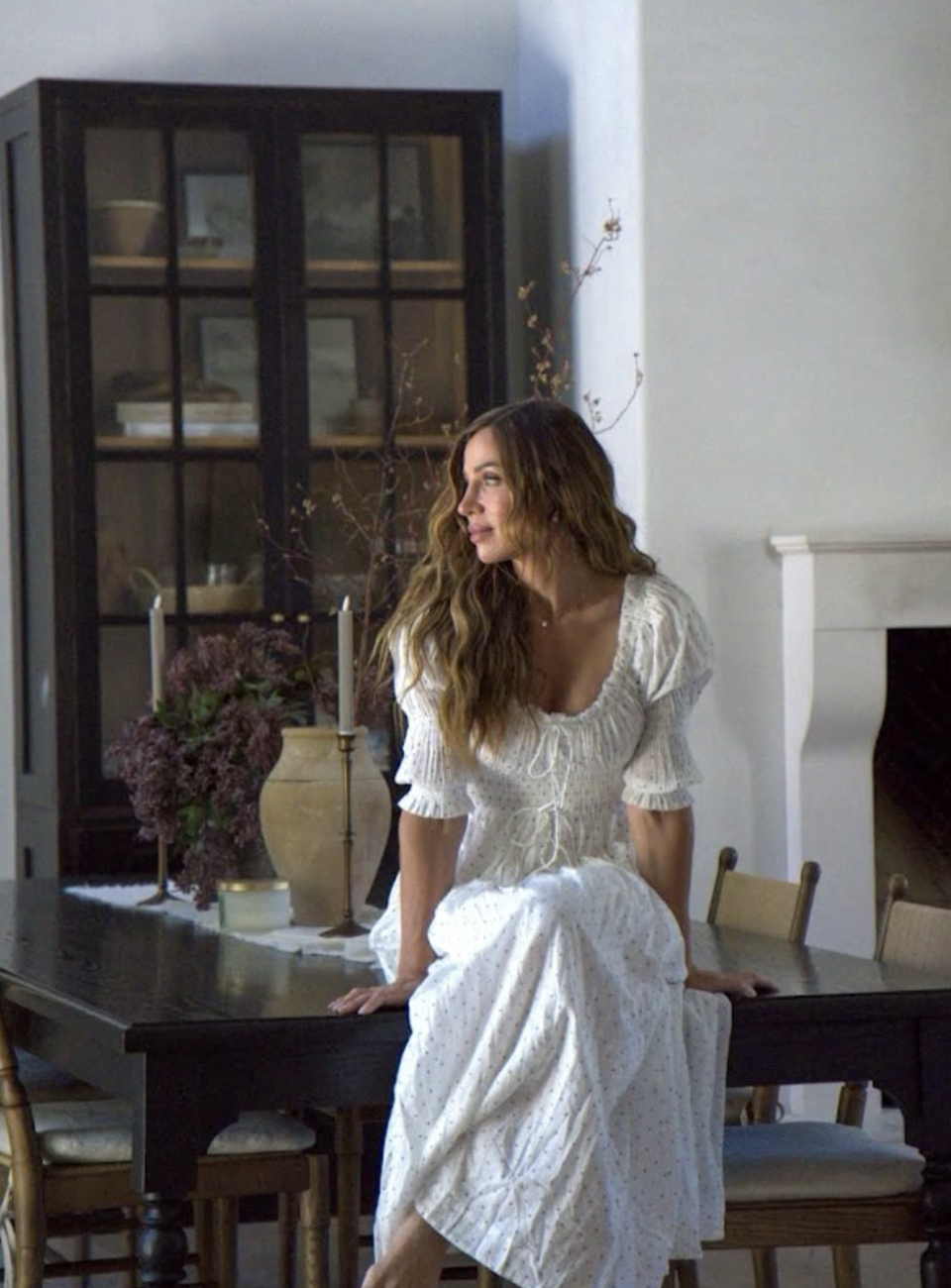A woman with wavy brown hair sitting on a dining table in a cozy room, wearing a white dress with a smocked bodice, surrounded by flowers and candles in a decorative setting.
