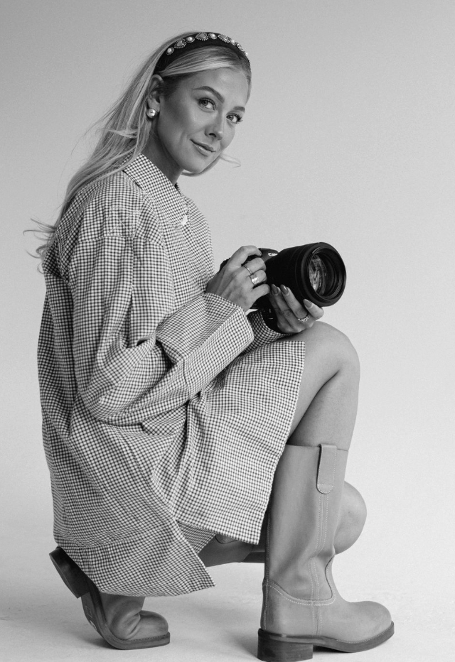 Black and white photo of a woman sitting on a stool, holding a camera, wearing a checkered dress, boots, earrings, and a headband, smiling at the camera.