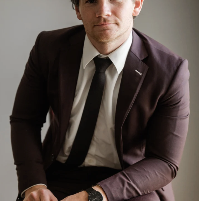 A man wearing a dark suit, white shirt, and black tie sitting against a gray background.
