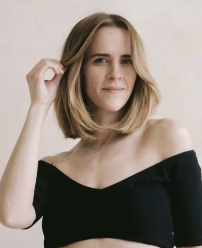 A woman with shoulder-length blonde hair, wearing an off-the-shoulder black top, looking at the camera with a slight smile, touching her hair with one hand.