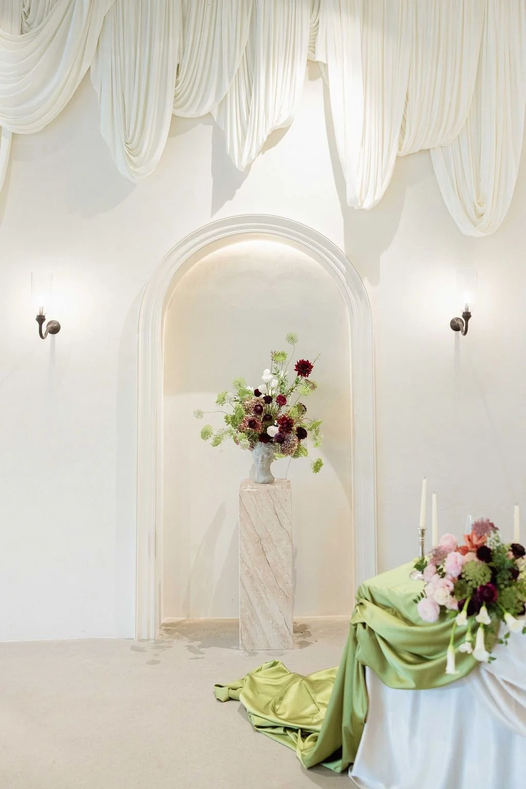Elegant white room with draped curtains, wall-mounted sconces, an arched wall niche with a floral arrangement on a marble pedestal, and a table with a floral centerpiece and candles draped with green fabric.