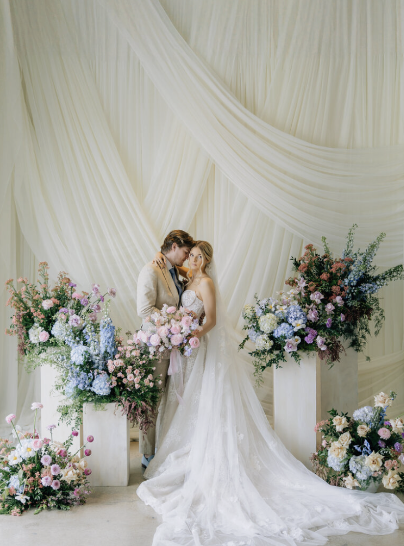 A bride and groom standing close together, surrounded by large arrangements of pink, purple, and white flowers, in front of white draped fabric at a wedding ceremony.