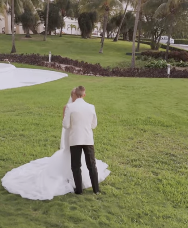 A couple dressed in wedding attire standing on a lush green lawn surrounded by trees and landscaping.