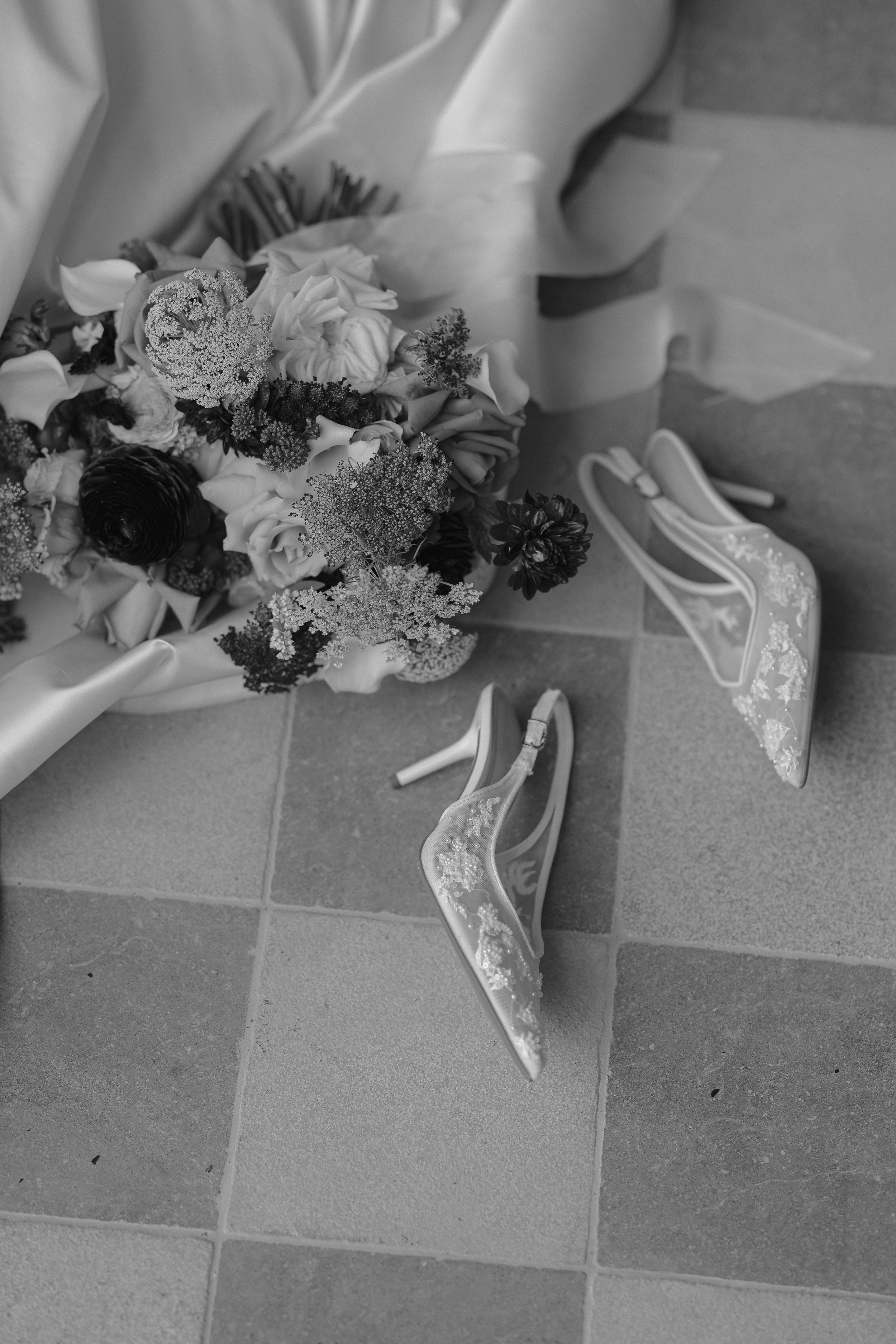 A bouquet of flowers lying on the floor near a pair of high-heeled shoes. The flowers include roses and other assorted blooms, wrapped in satin ribbon, placed on a tiled floor.