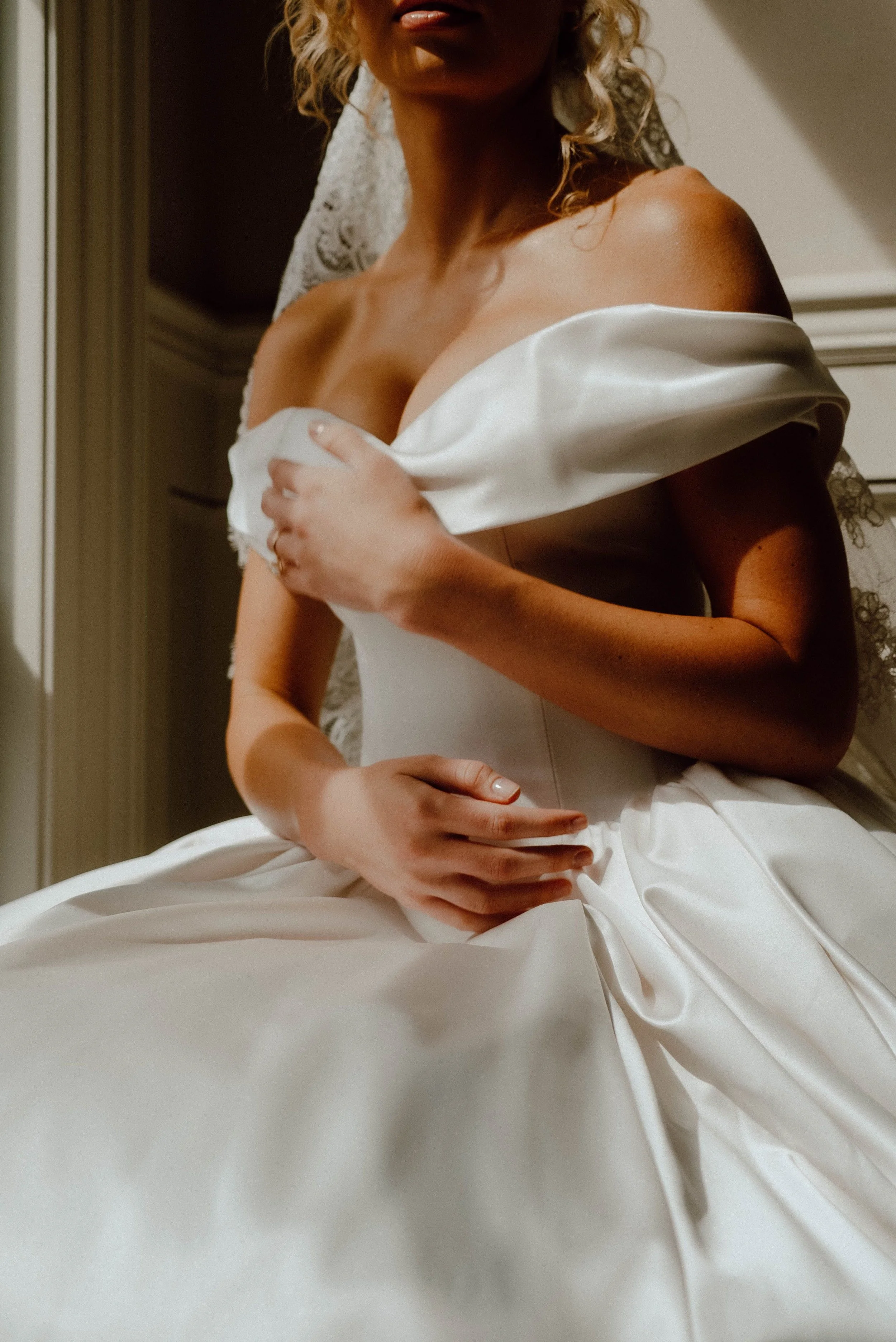 A woman in a wedding dress touches her shoulder and chest with her hands, with a lace veil visible in the background.