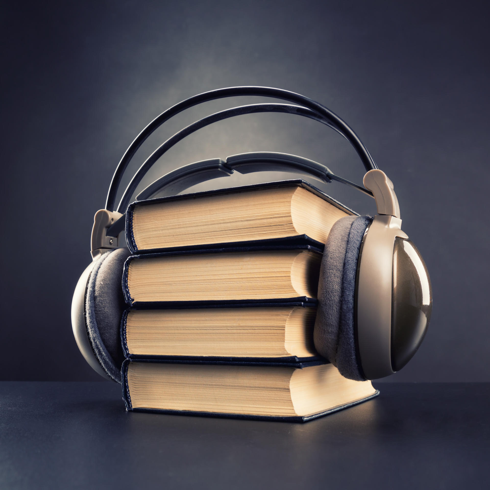Stacked hardcover books with a pair of over-ear headphones placed on top, on a dark surface with a dark background.