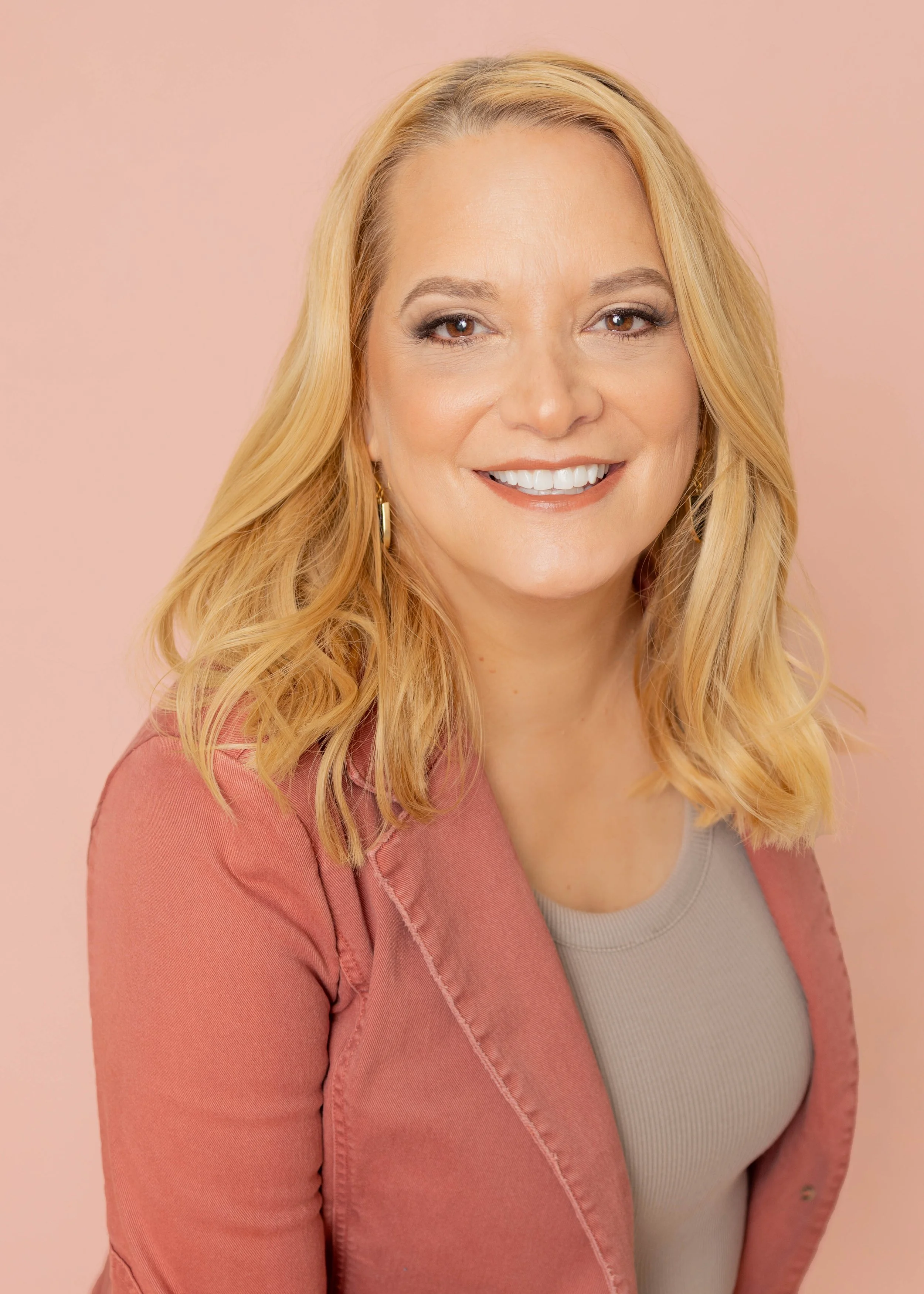 A woman with blonde, wavy hair smiling in front of a light pink background, wearing a pink blazer over a beige top.