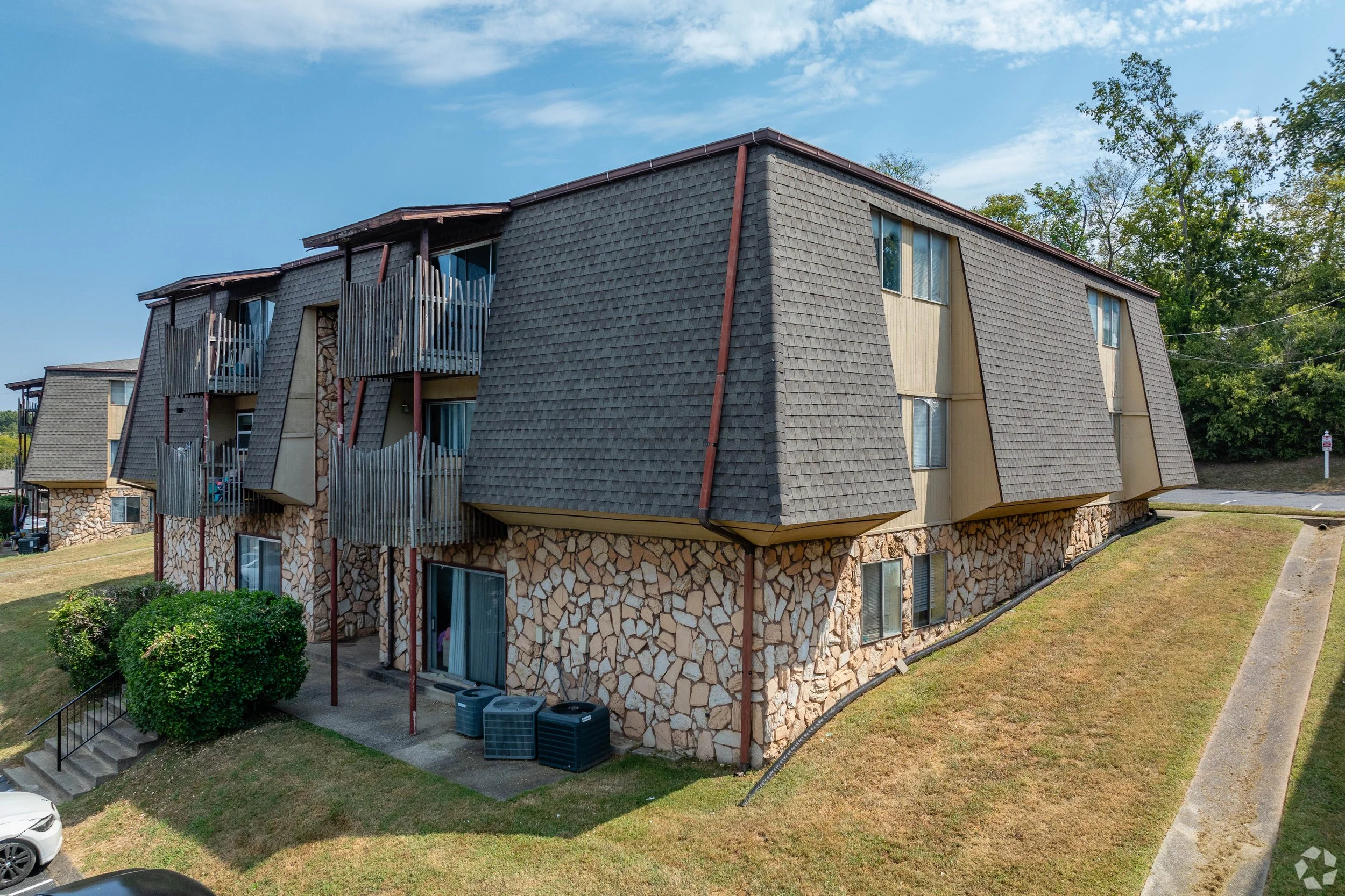 Multi-story residential building with stone and wood exterior, steep roof, and small balconies, set amidst green lawns and trees under a partly cloudy sky.