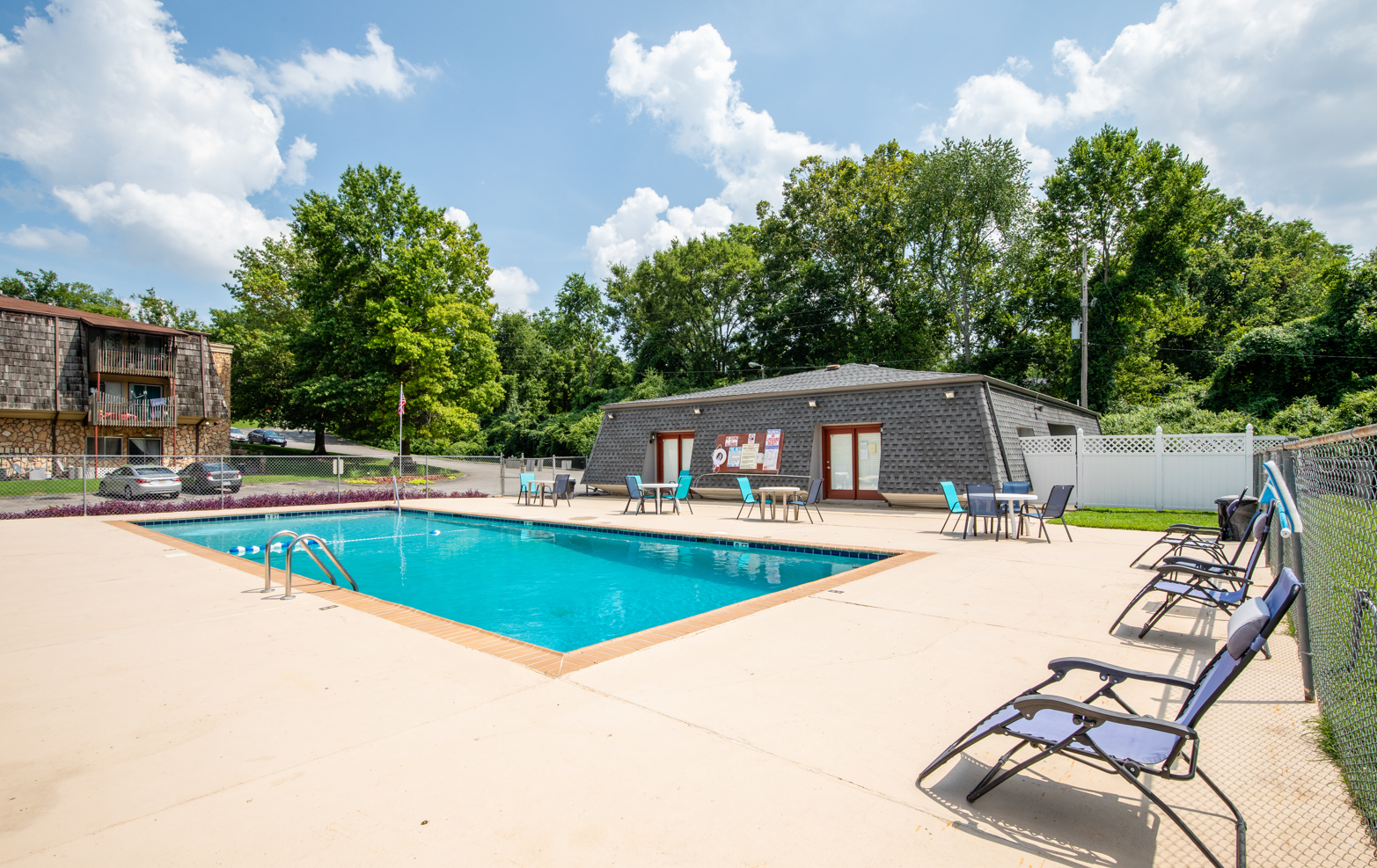 Outdoor swimming pool with lounge chairs, tables, and a building with bulletin board, surrounded by trees and clear blue sky.