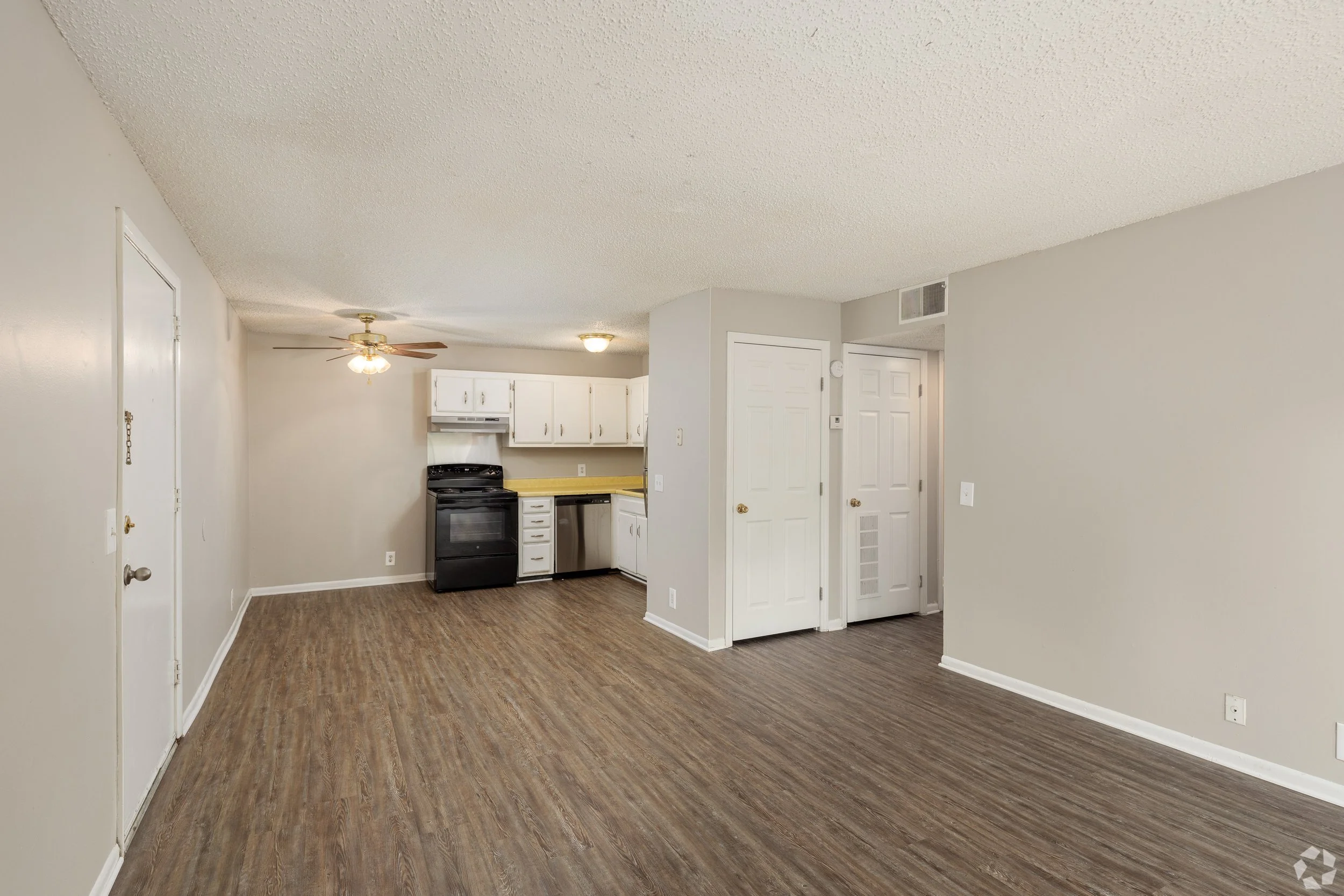 An open living space with wood-look flooring, beige walls, a kitchen with white cabinets, a black stove, and a ceiling fan with a light, viewed from the dining area.