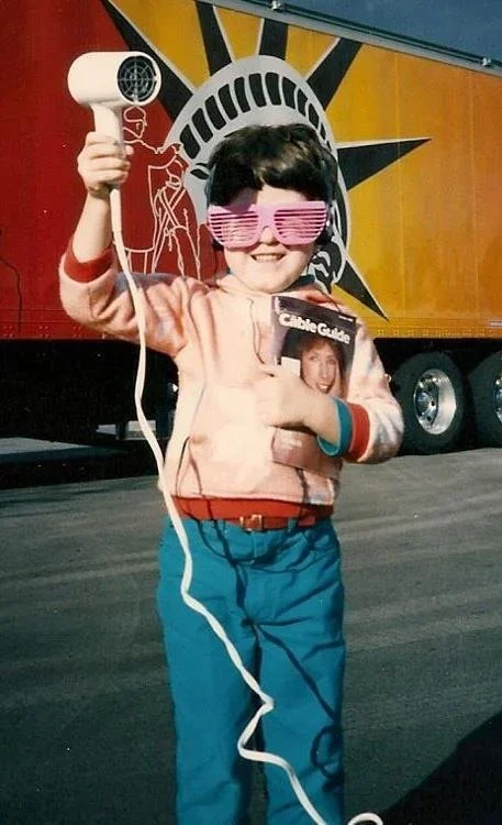 A young boy stands in front of a trailer with New York City and the Statue of Liberty logo, wearing pink shutter sunglasses, holding a white landline phone and a magazine, smiling cheerfully.