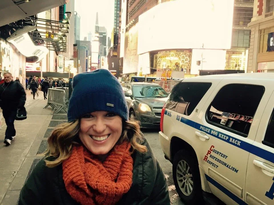 A smiling woman wearing a navy beanie and a red scarf stands on a busy city sidewalk with tall buildings and a police vehicle in the background.
