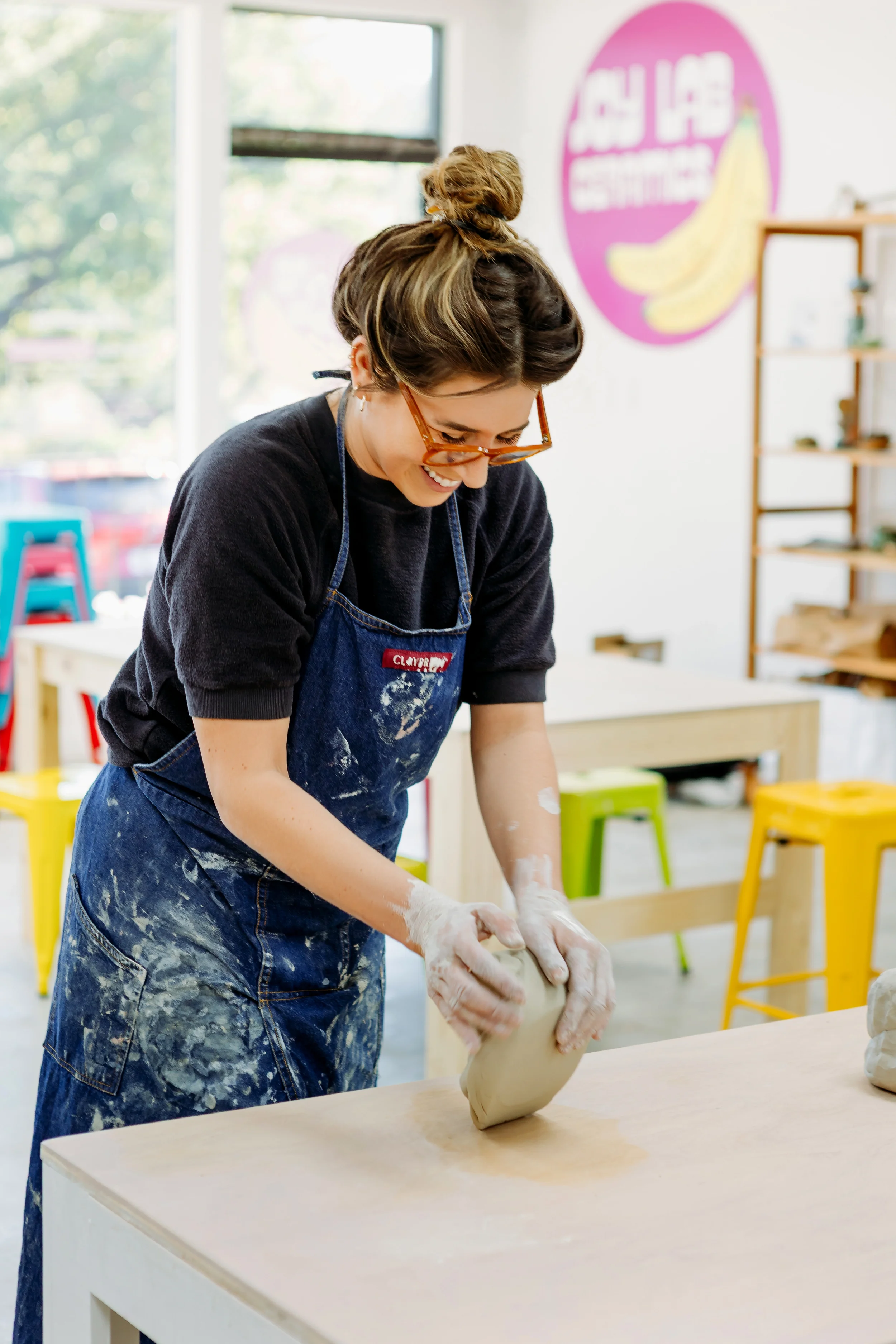 A woman with glasses, wearing a black shirt and apron, shaping clay on a table in a brightly lit art studio.