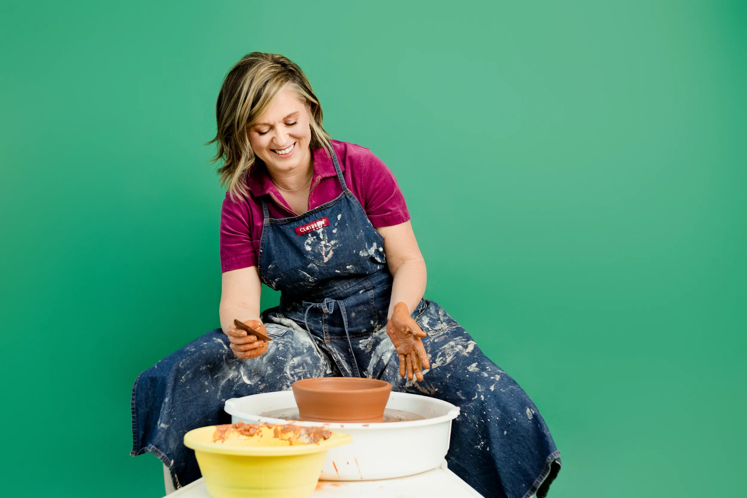 A woman smiling while shaping clay on a pottery wheel, wearing a pink shirt, denim apron, and paint-splattered jeans against a green background.