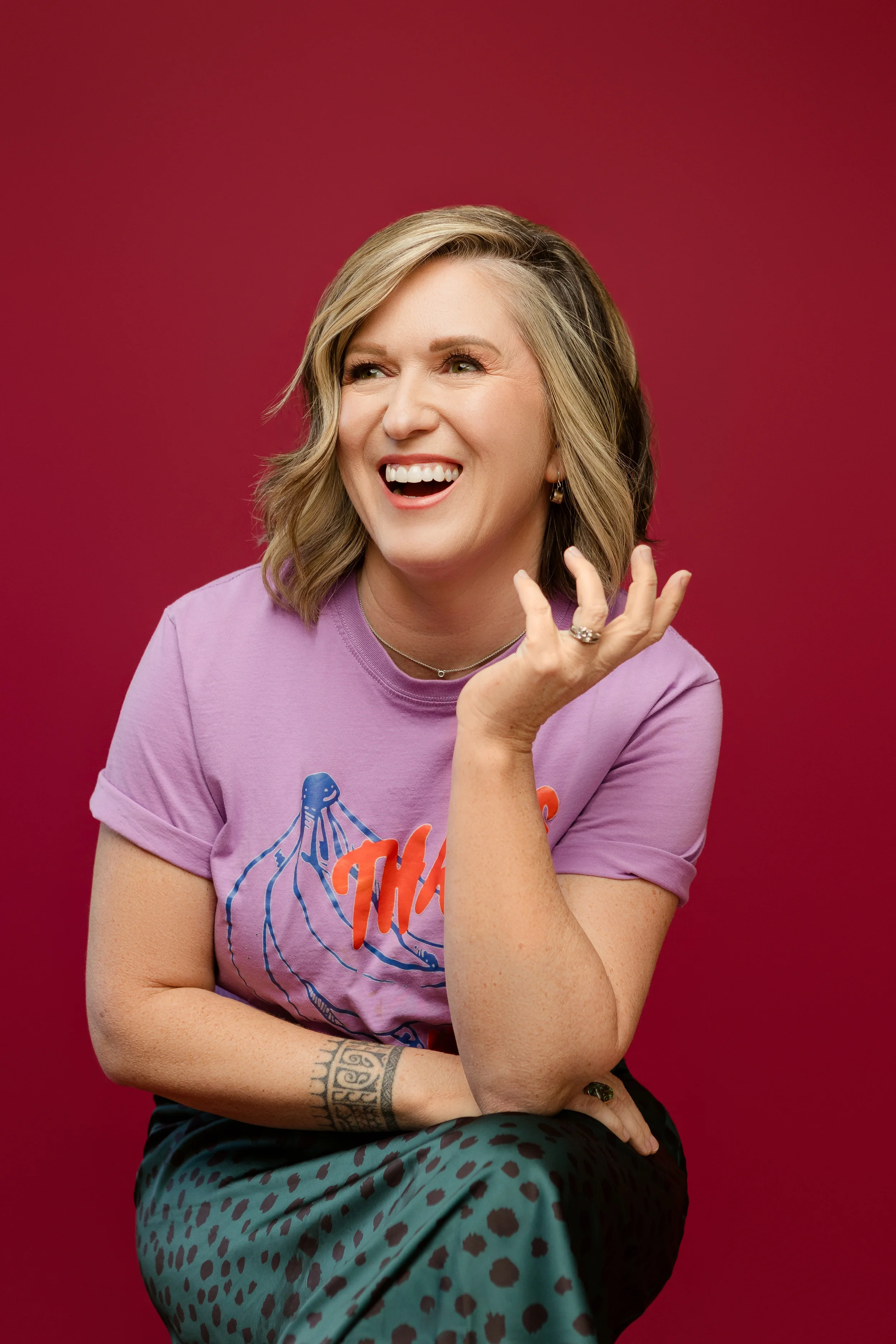 Woman with blonde hair wearing a purple t-shirt with graphic print, sitting against a red background, smiling, with one hand near her face and tattoos visible on her arm.