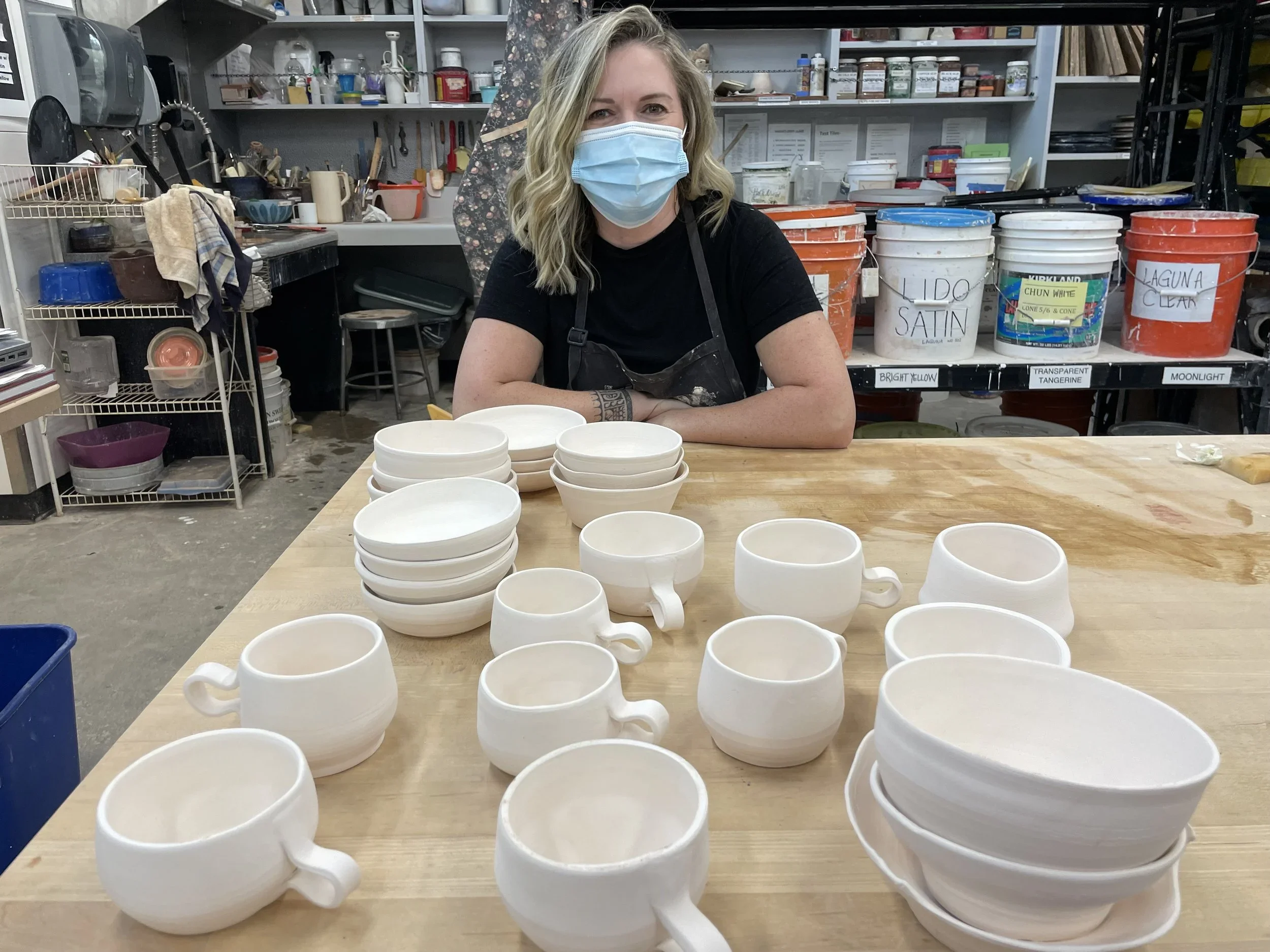 A woman wearing a mask leaning on a table with white ceramic cups, in a pottery studio with shelves of paint buckets and tools in the background.