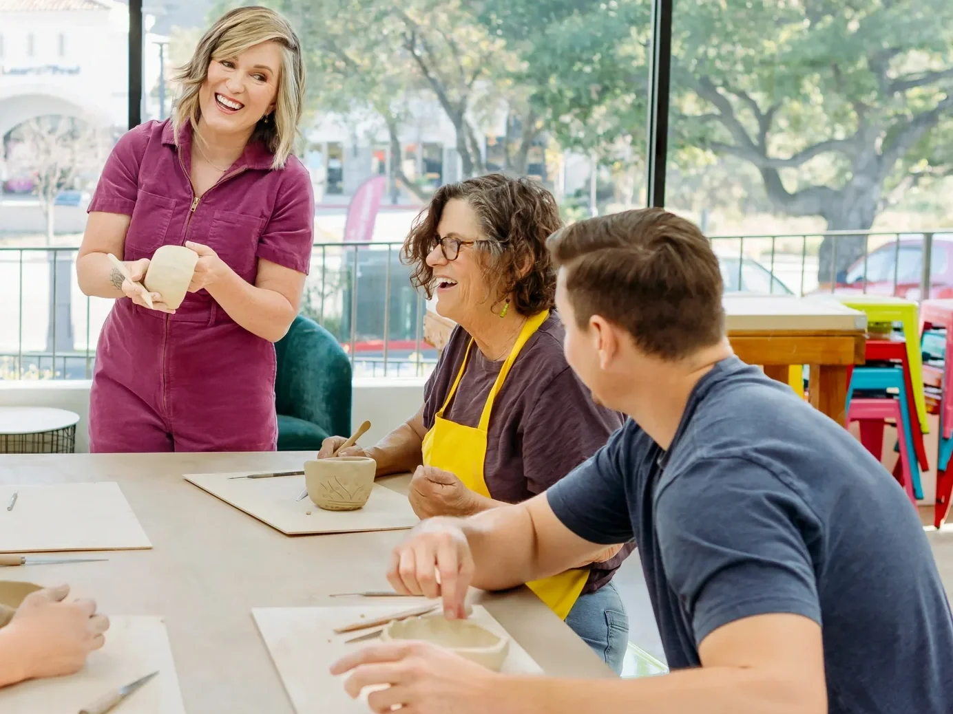 A woman demonstrating pottery to a group of people in a bright room with large windows and a view of trees and parked cars outside.