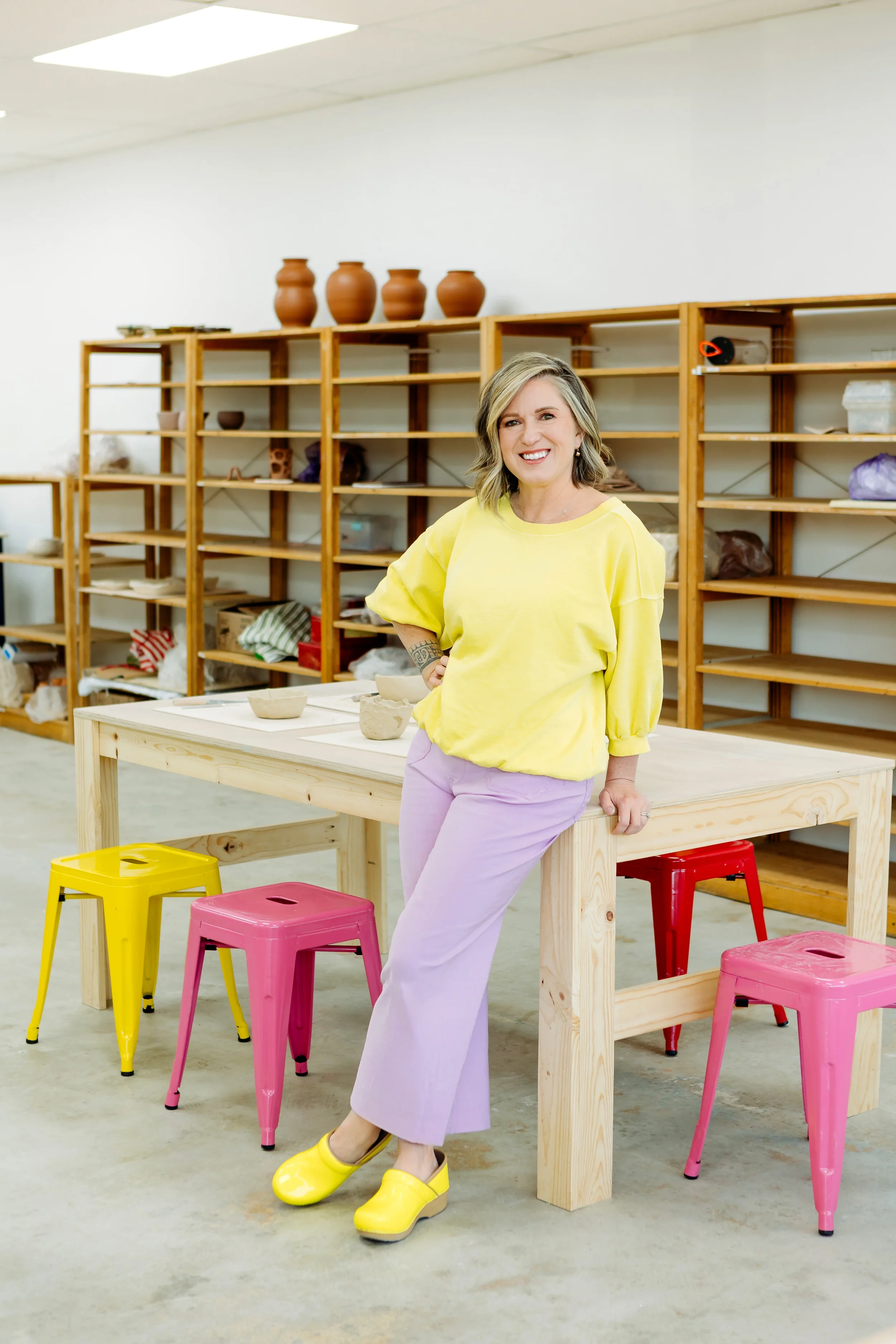 A woman standing in a pottery studio, smiling, wearing a yellow sweatshirt, light purple pants, and yellow shoes. The studio has wooden shelves with pottery and supplies, a wooden table with pottery pieces on it, and colorful plastic stools.