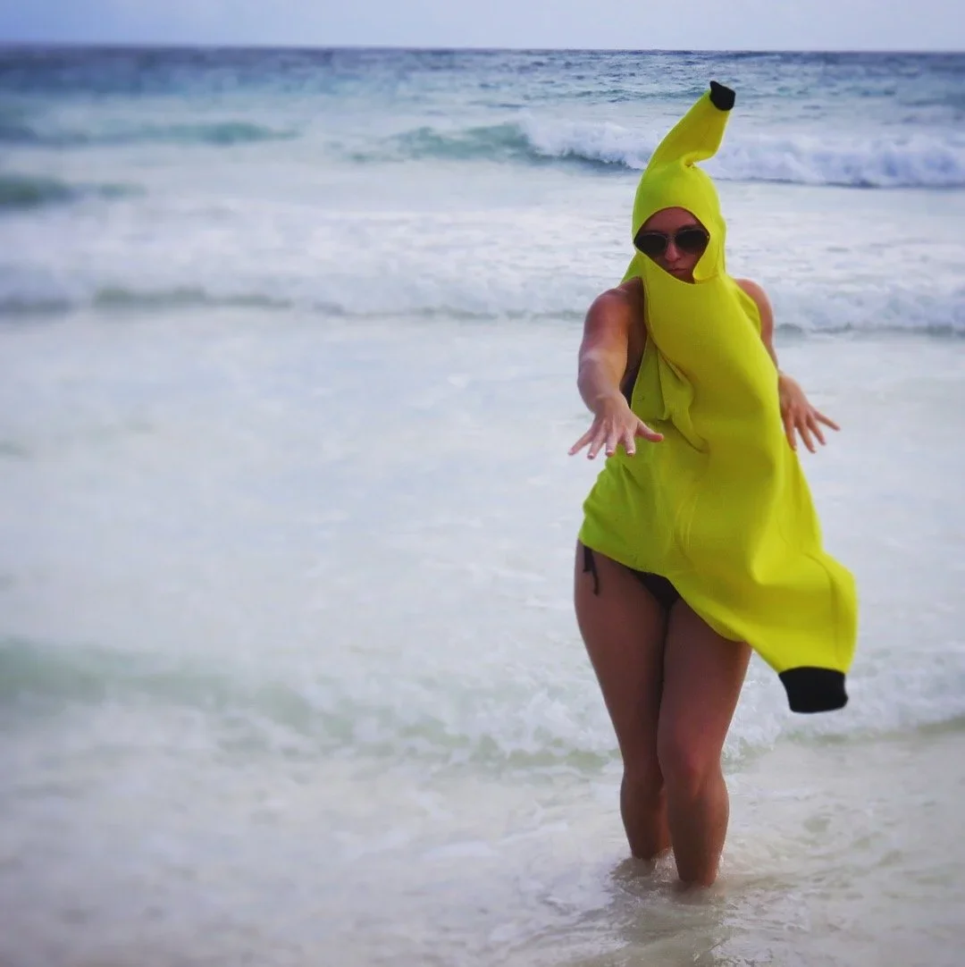 Person on the beach wearing a bright yellow banana costume with sunglasses, reaching out with both hands.