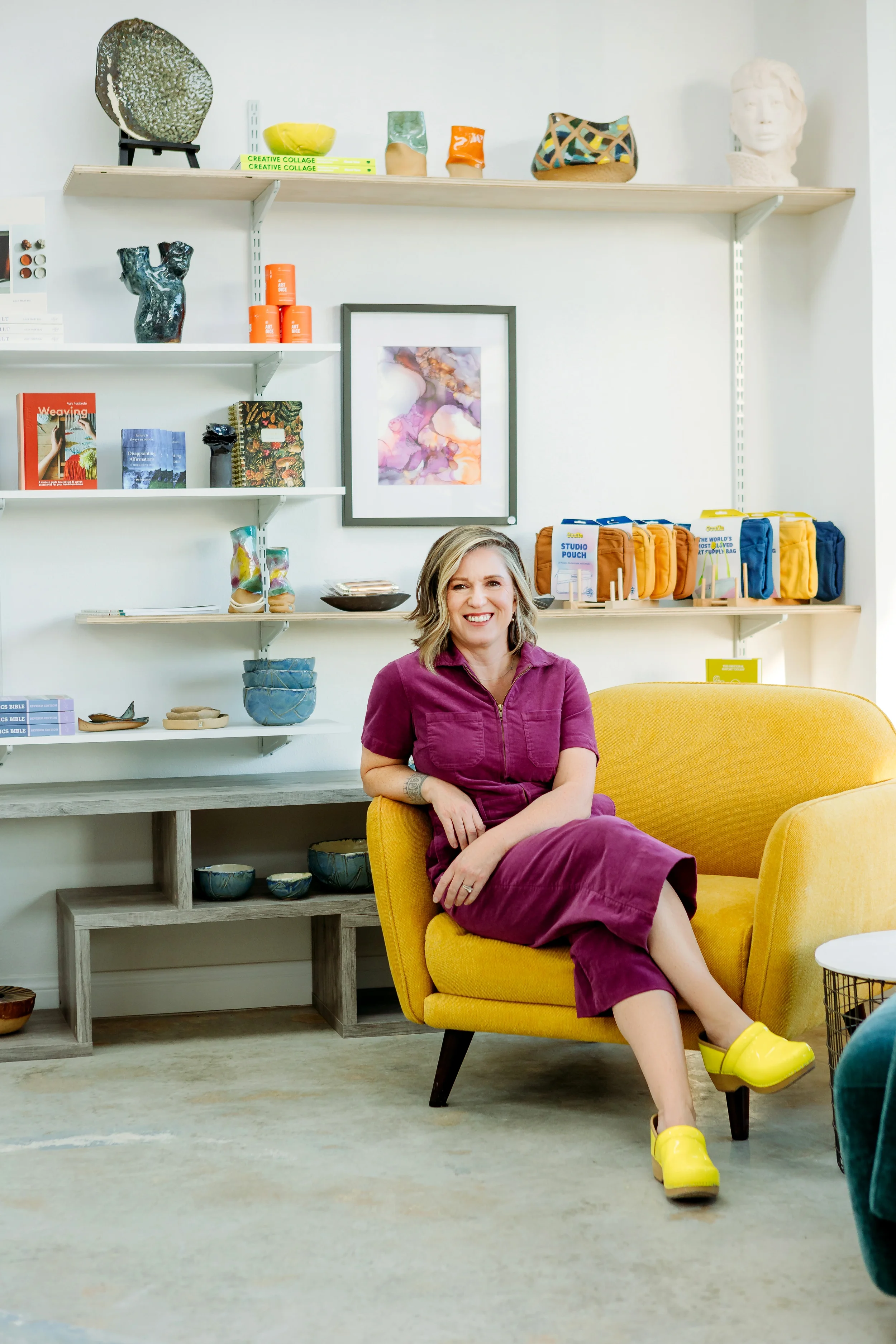 A smiling woman sitting in a yellow armchair in a room with shelves holding decor items, books, and pottery. She is wearing a magenta dress and bright yellow shoes.
