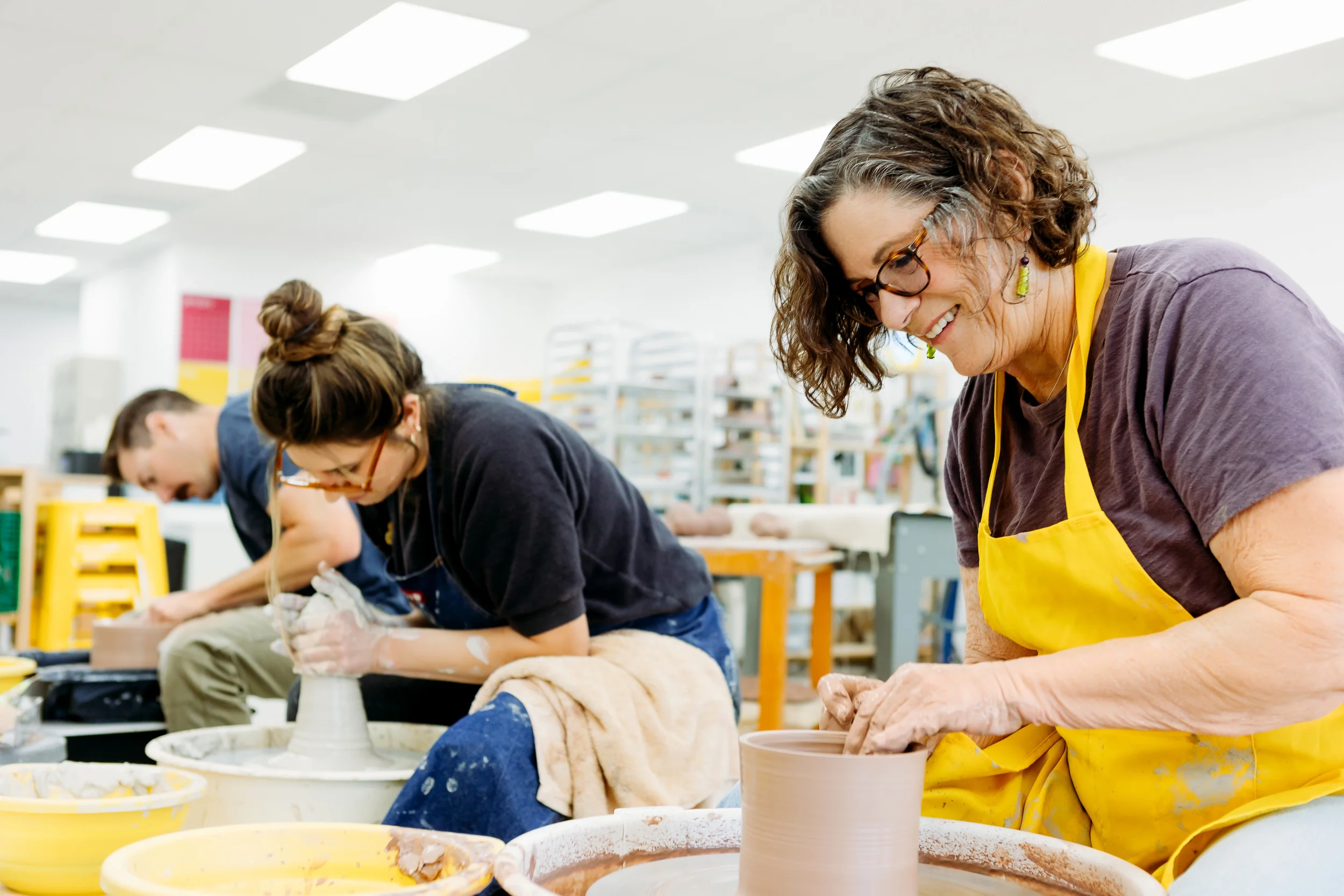 Women working on pottery in a ceramics classroom, with focus on a woman wearing glasses and a yellow apron, smiling as she shapes clay on a pottery wheel.
