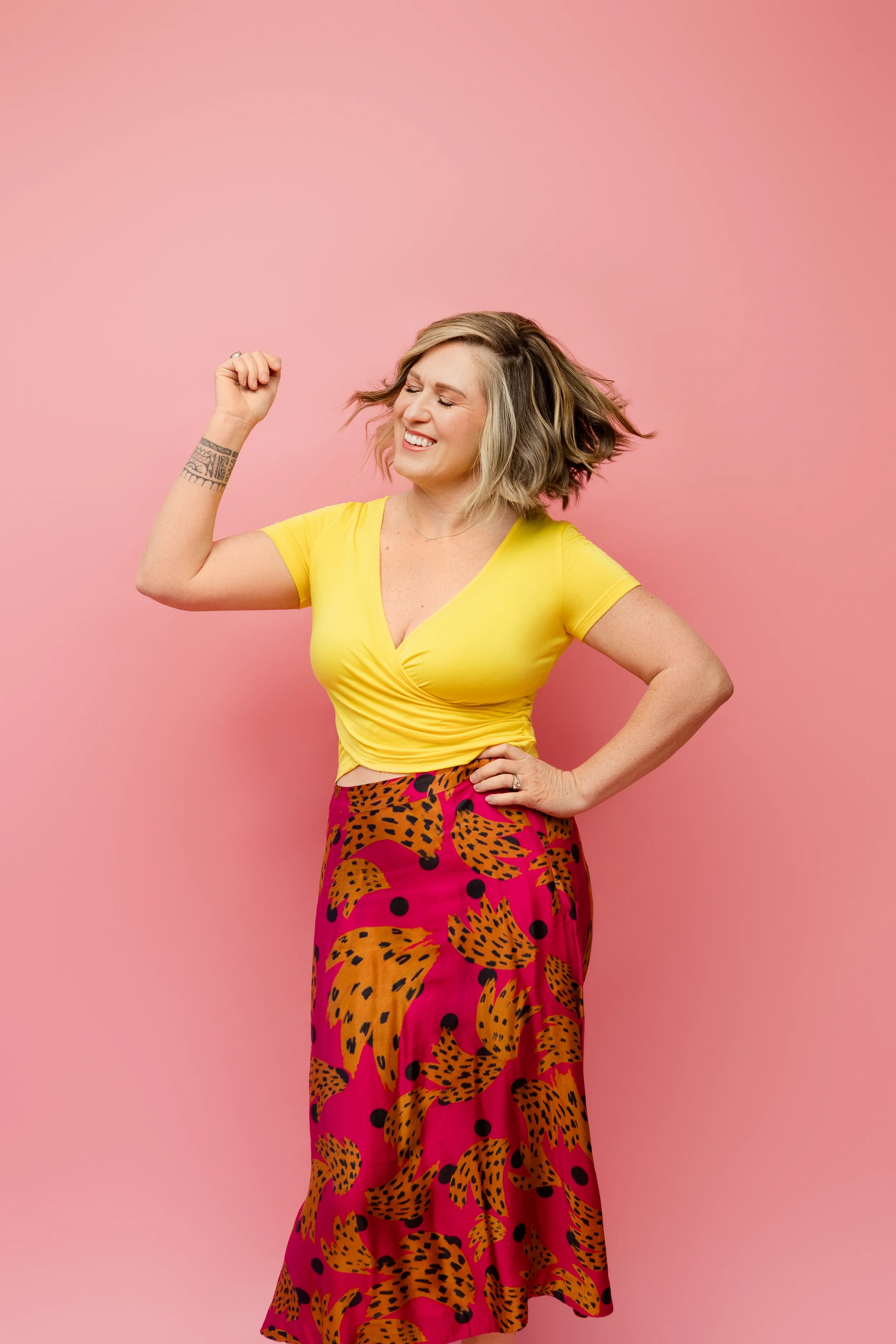 A woman with short wavy blonde hair wearing a yellow top and a long pink skirt with black and orange leopard print pattern, standing against a pink background. She is smiling and dancing with her eyes closed, her right arm raised and her left hand on her hip.