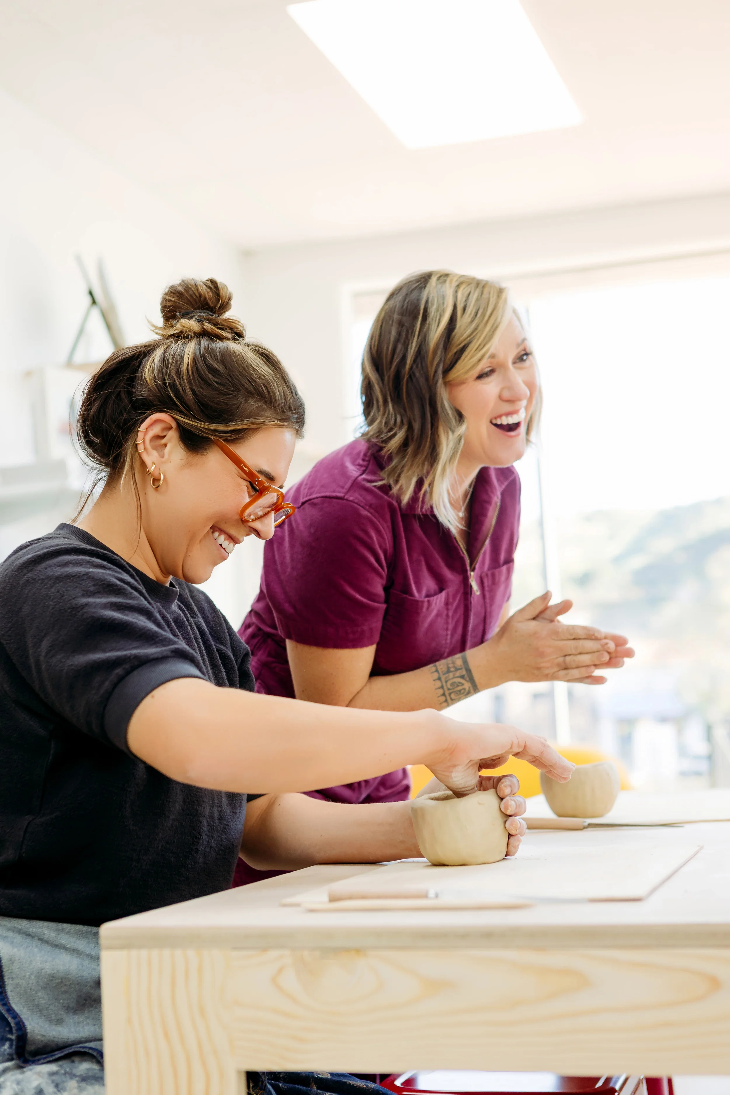 Two women smiling and engaging in pottery class, shaping clay bowls on a wooden table indoors with natural light.