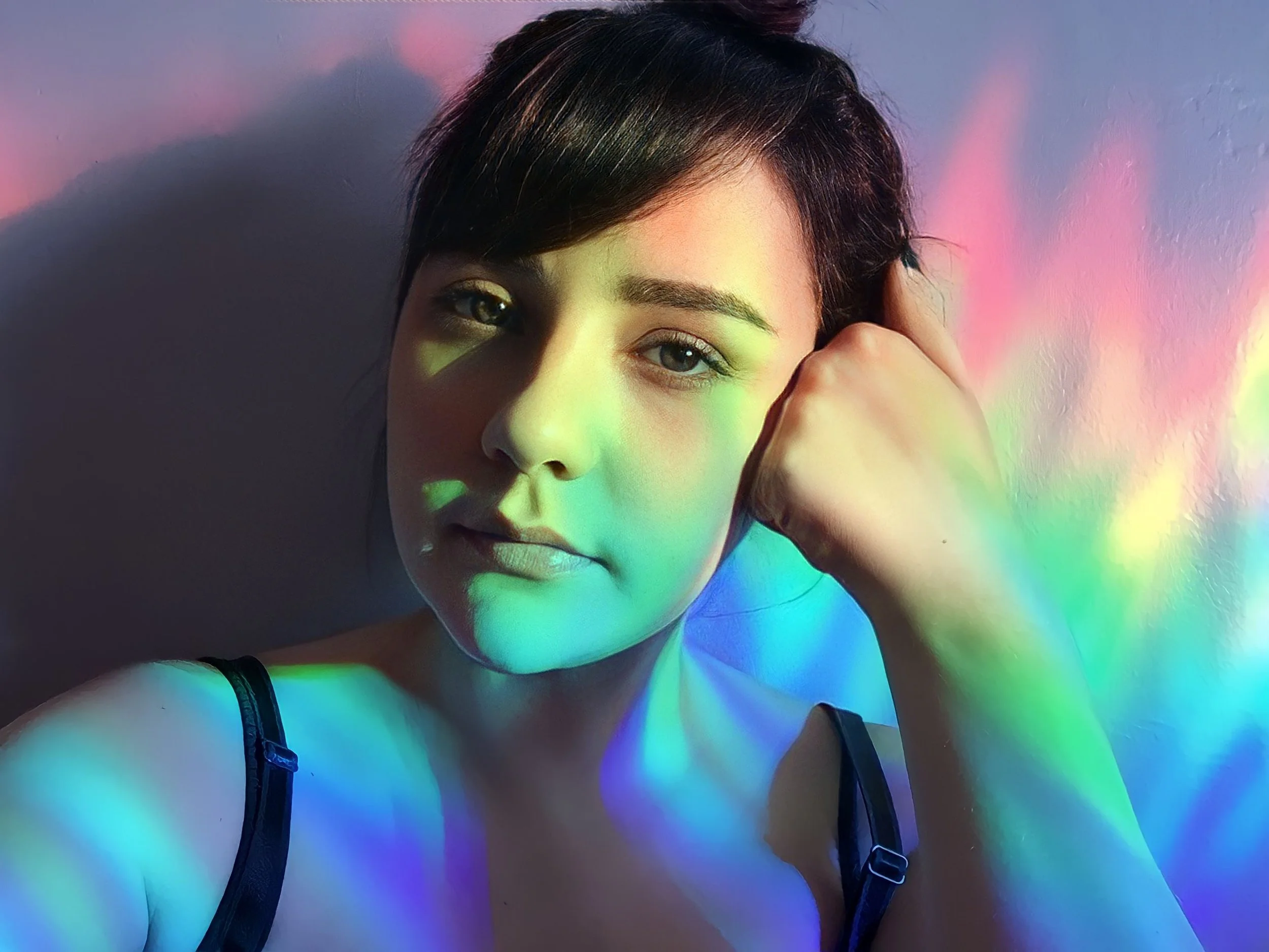 A woman with short dark hair, blue eyes, and light skin, holding her head with her right hand, in front of a colorful rainbow projected on a wall.