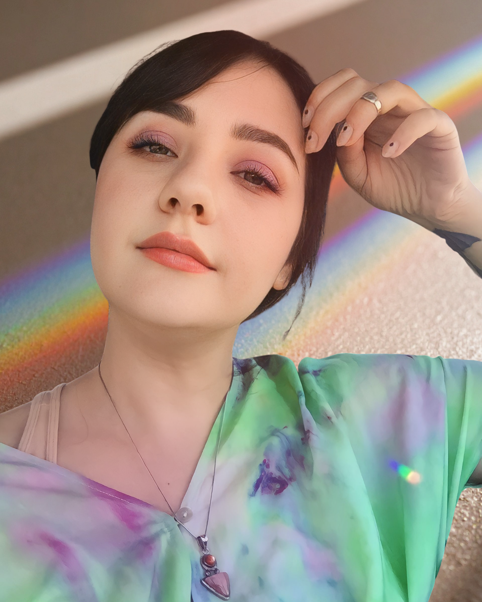 Close-up of a woman with short dark hair, wearing pink eyeshadow and a green tie-dye shirt, holding her hand up to her head, with a rainbow reflection background.